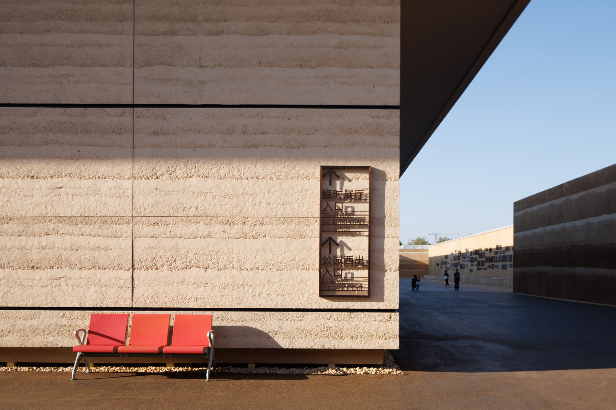 Travertine wall detail with recessed bronze panel above a red bench in sharp shadow
