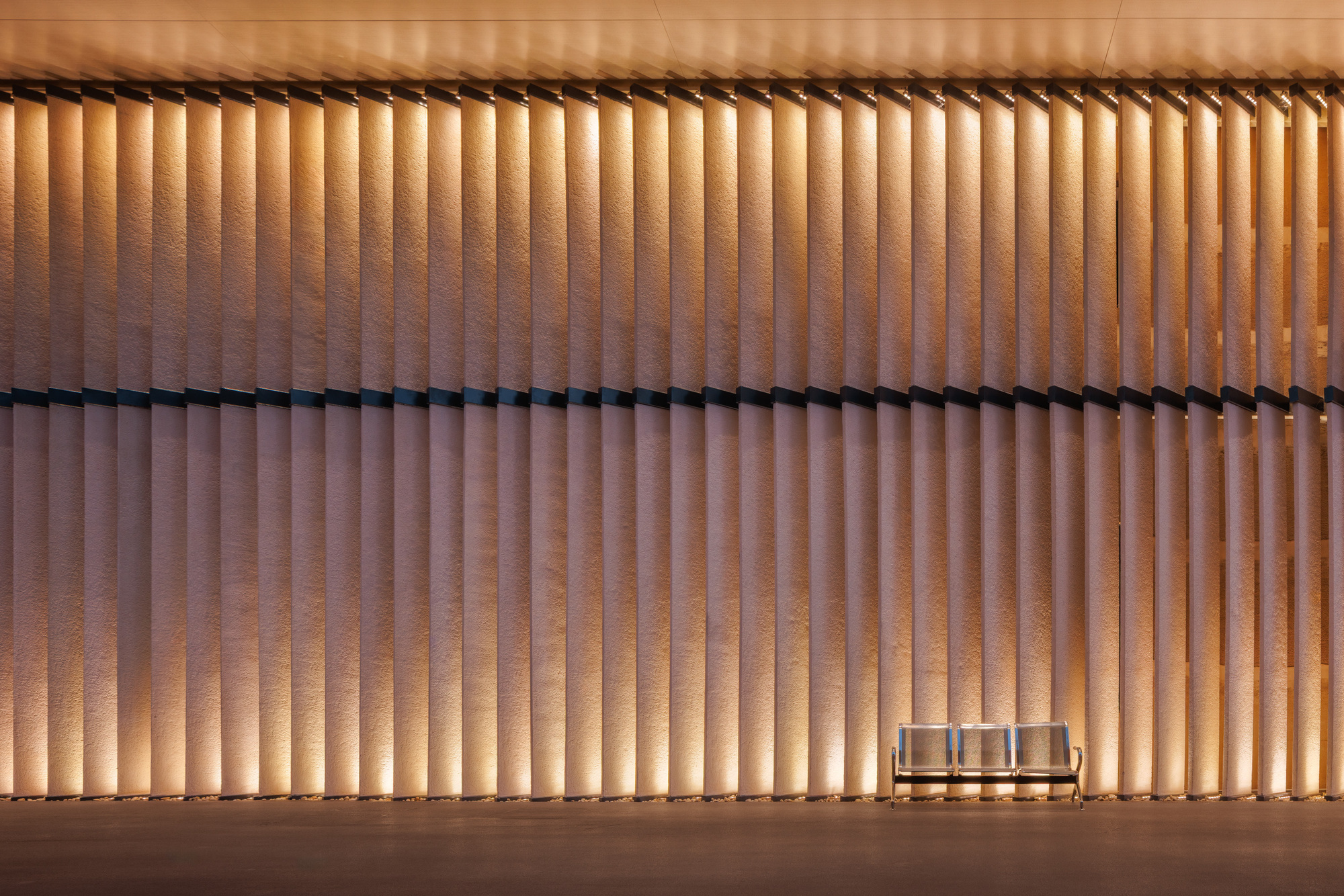 Vertical timber screen facade with backlighting and small bench seating below at twilight