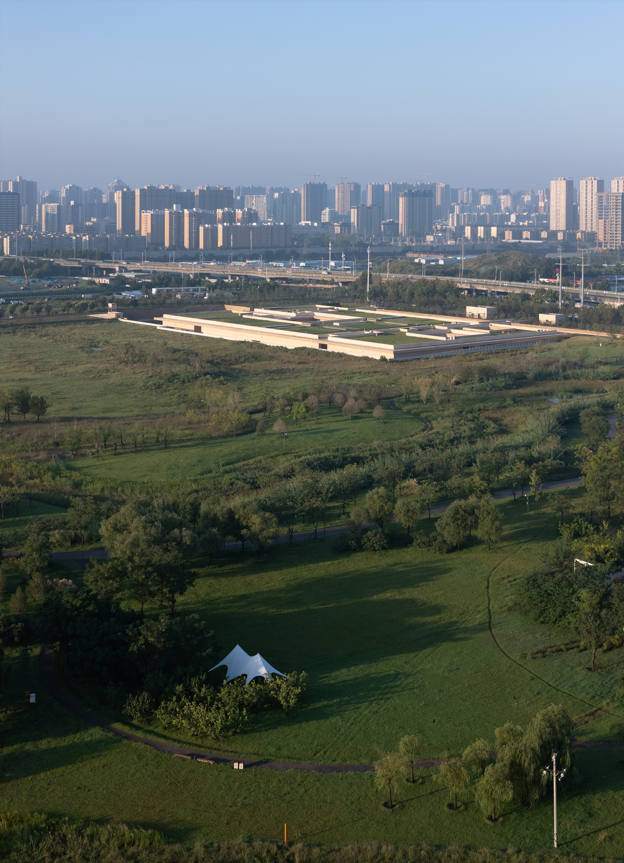 Aerial view of the low-slung building set within green parkland with a dense urban skyline beyond