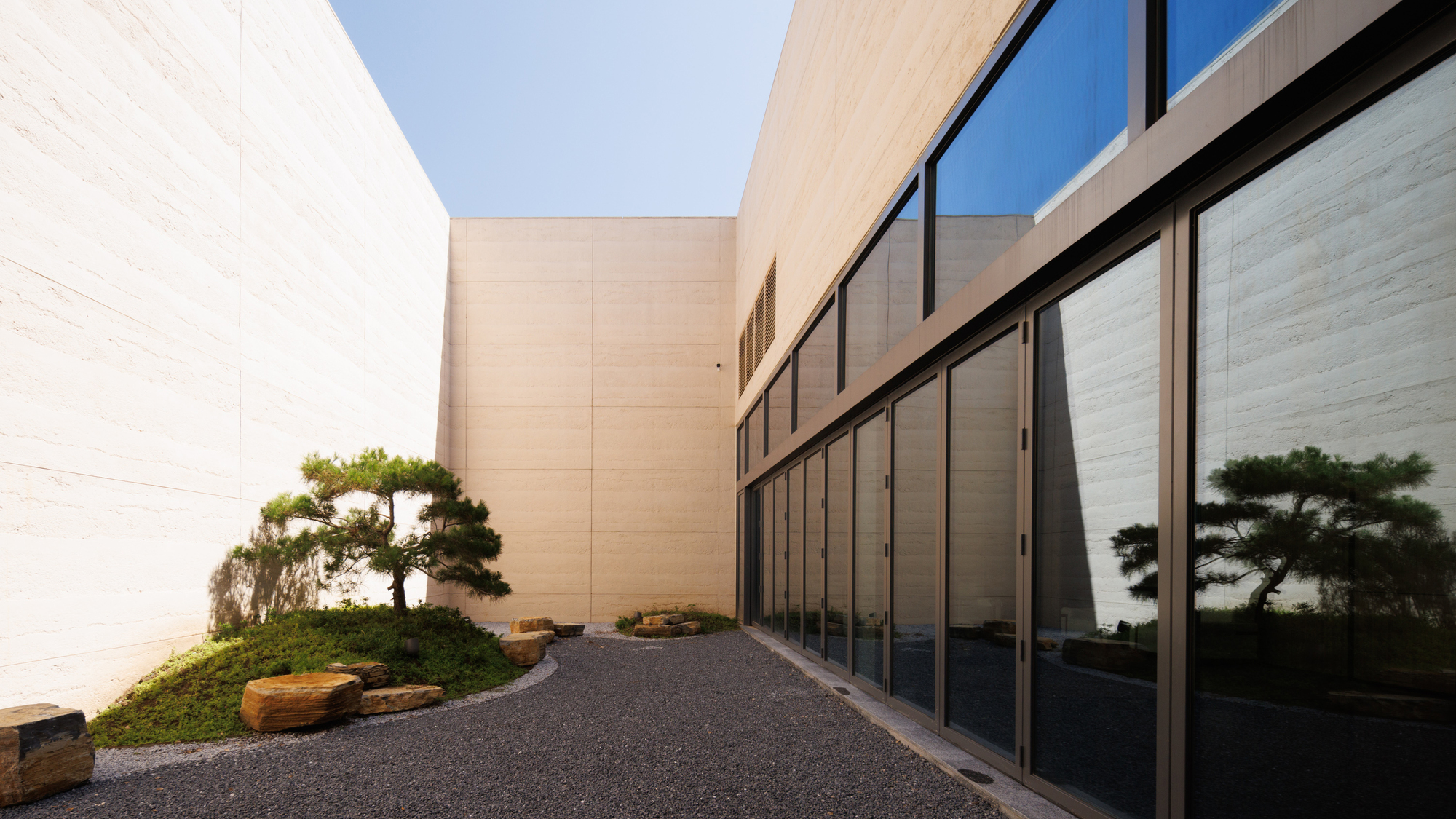 Courtyard with pale stone walls, floor-to-ceiling glazing and planted beds with sculpted pines