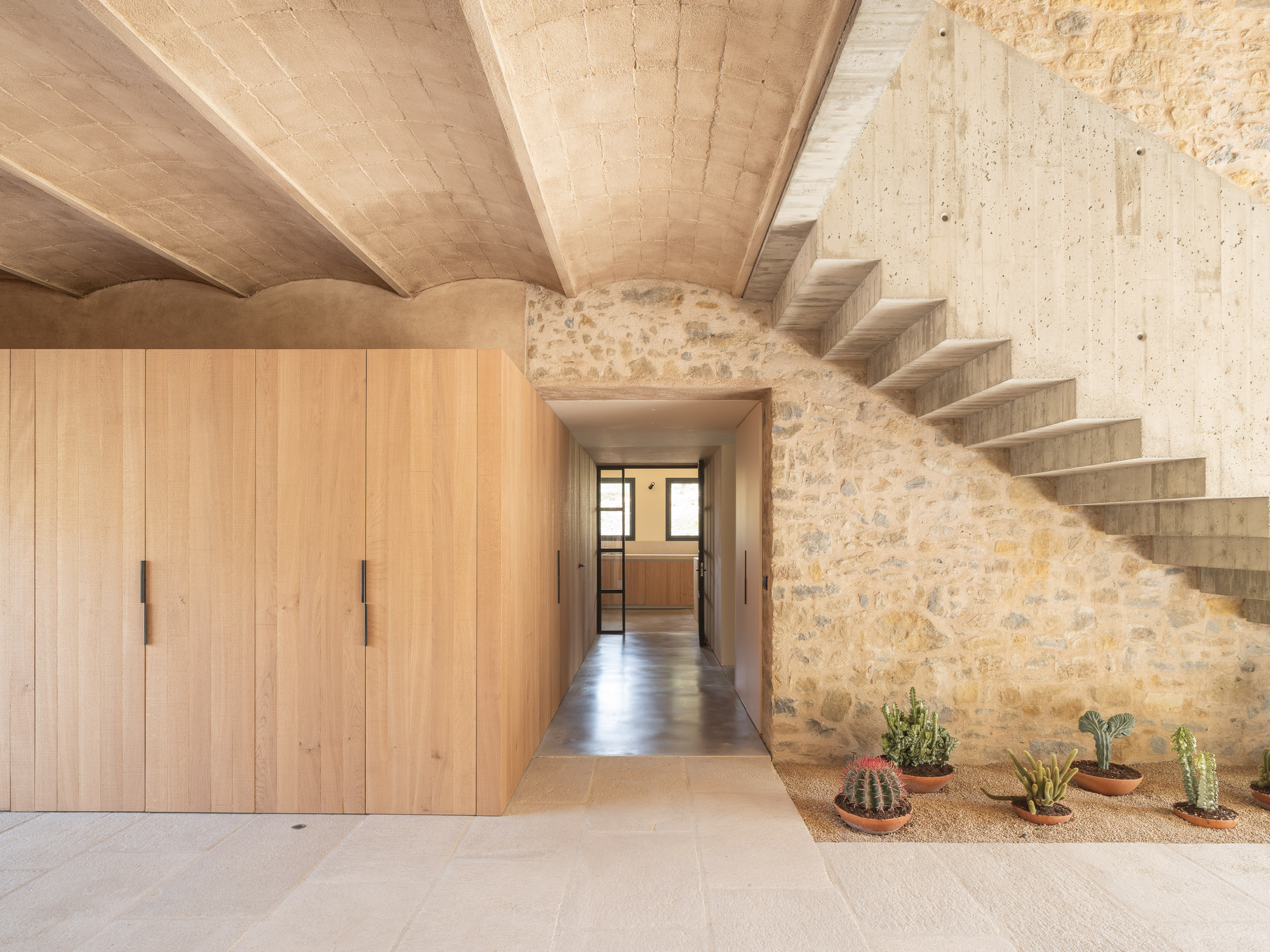 Concrete staircase rising along exposed stone wall with timber ceiling and potted cacti below