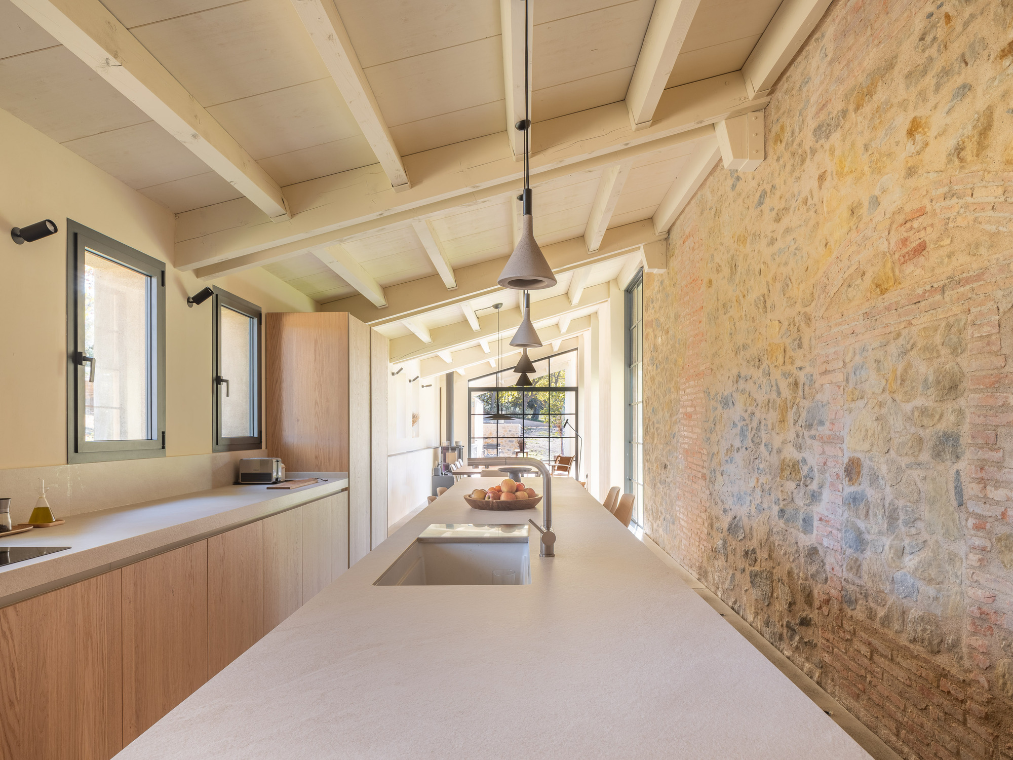 Kitchen with exposed timber beams and stone wall alongside light wood cabinetry and pendant lights
