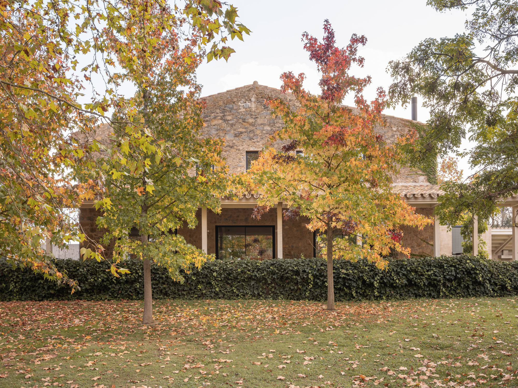 Stone volume with terracotta roof seen through deciduous trees shedding autumn leaves on the lawn