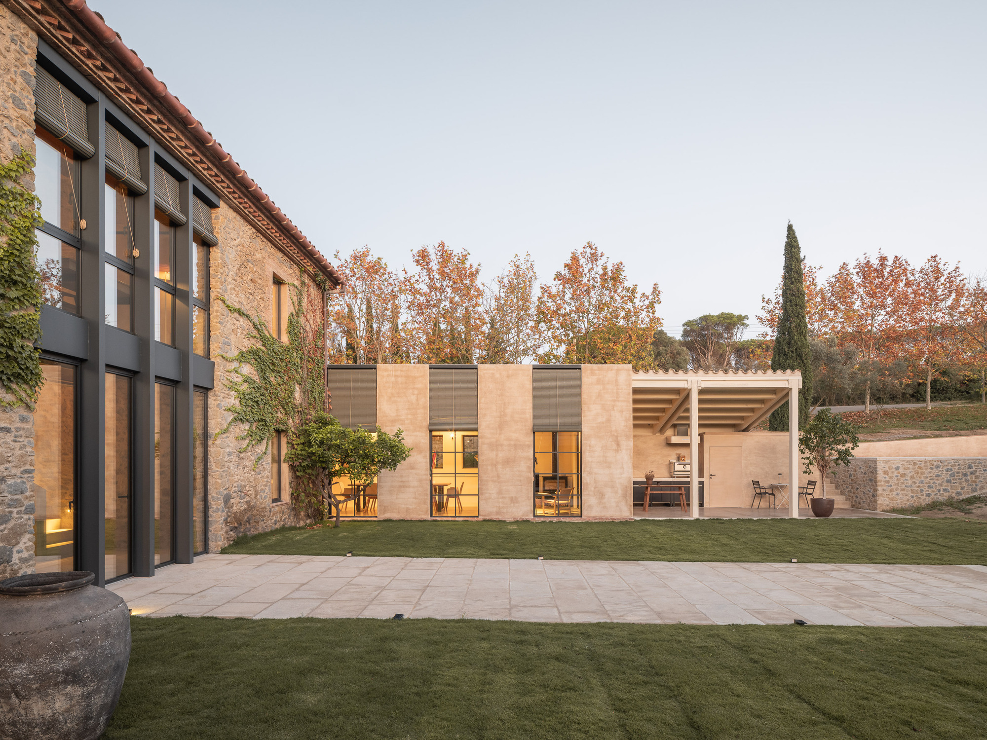 Garden view at dusk showing stone addition with alternating sandstone and glass panels beside terracotta-roofed volume