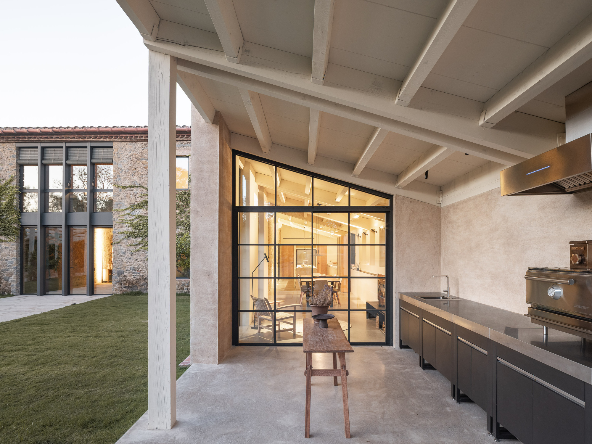 Covered outdoor kitchen with exposed beam ceiling and black-framed glass doors opening to an interior dining area