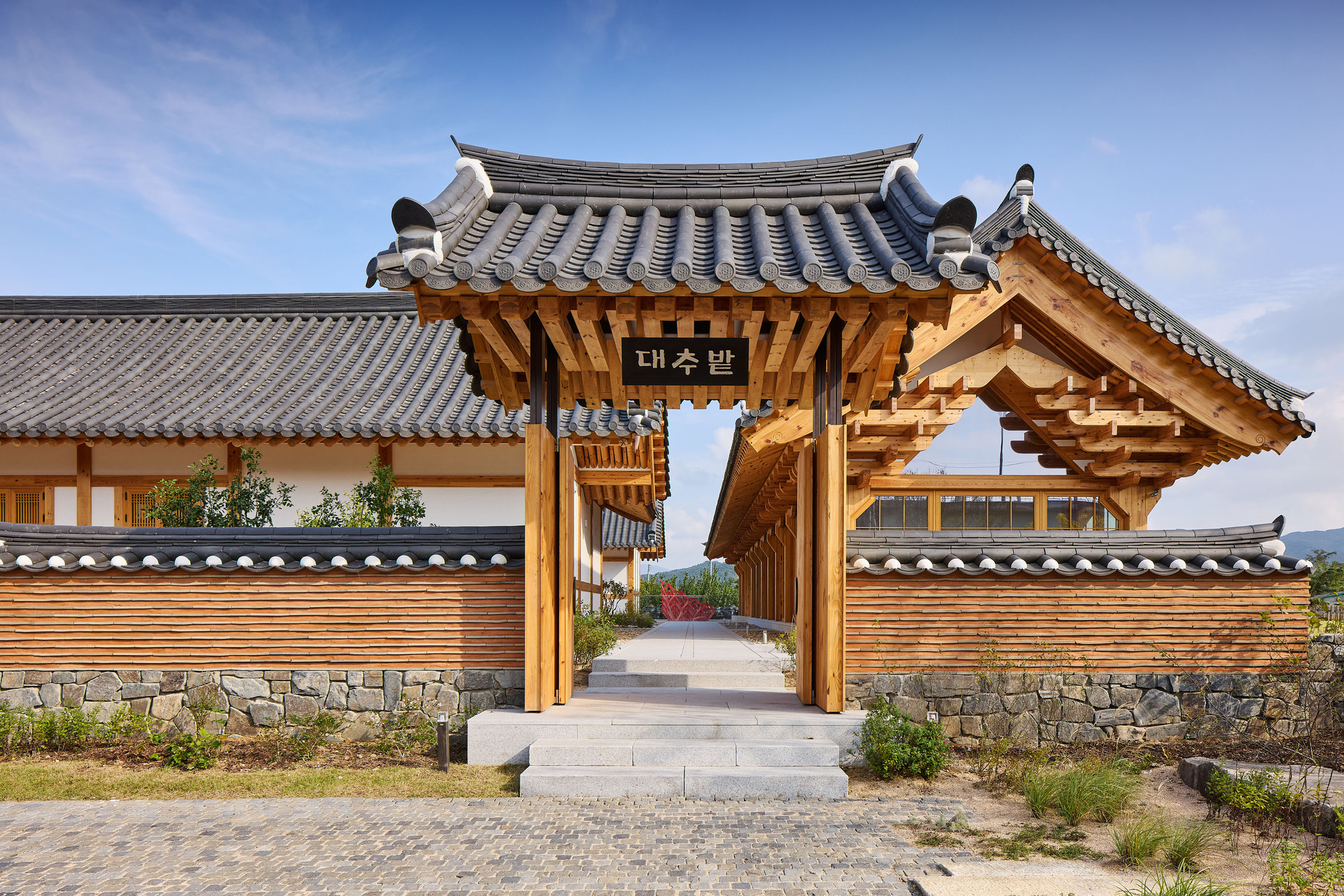 Timber entrance gate with curved tile roof and stone base wall under a clear blue sky