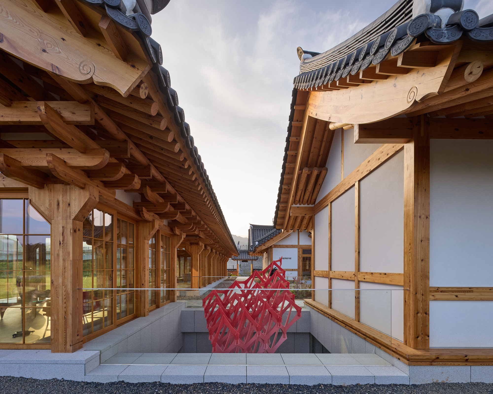 Sunken courtyard passage flanked by timber structures with a red sculptural installation at its center