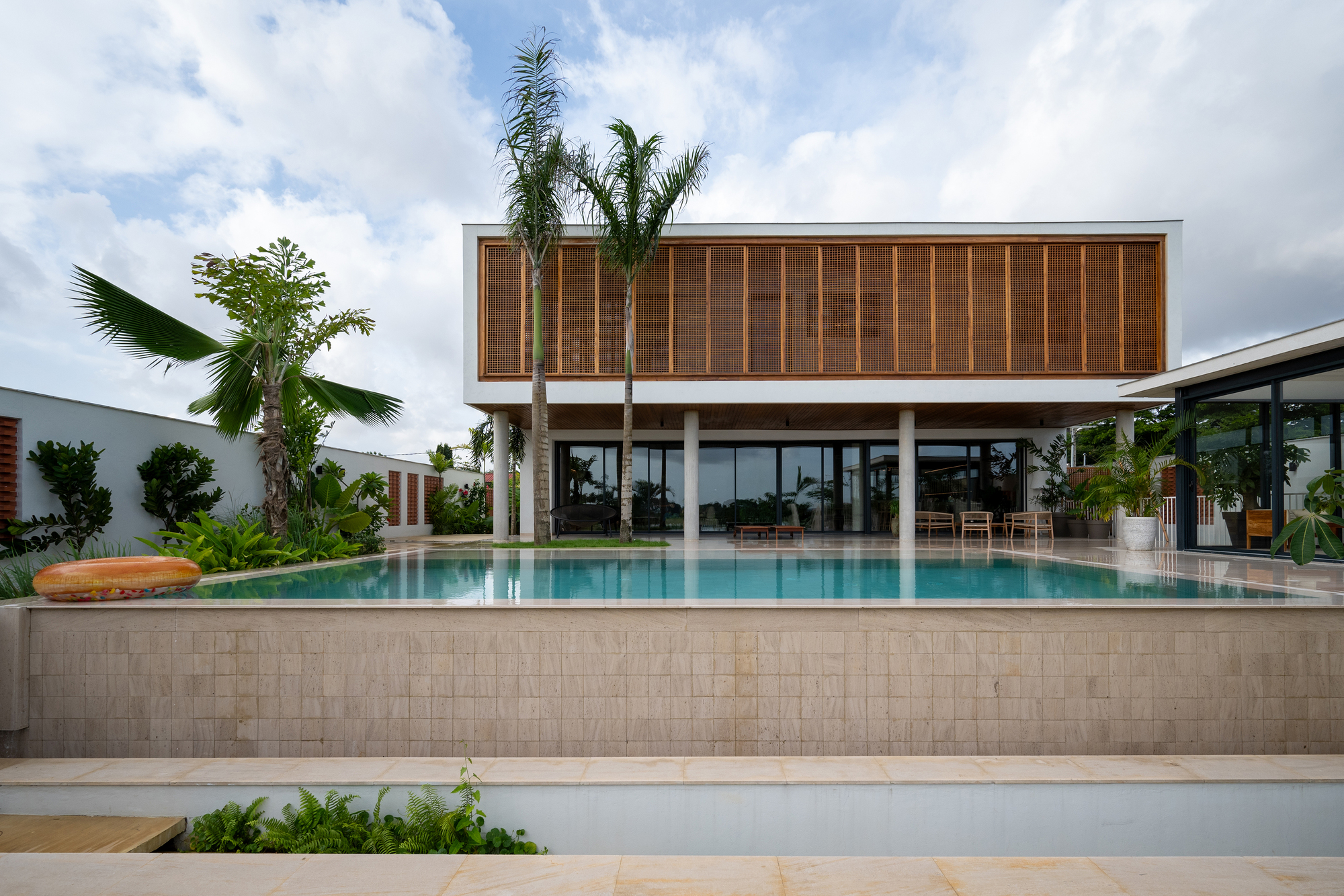 Elevated infinity pool with travertine tile surround facing a two-story volume with vertical timber louvers under cloudy skies