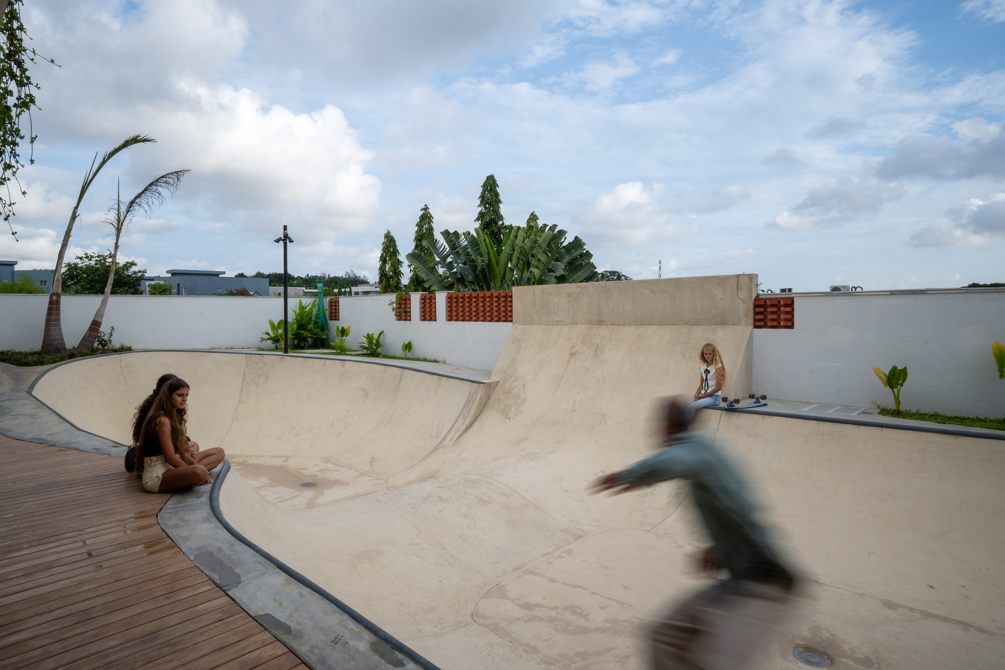 Concrete skateable bowl with two people seated and one in motion under a partly cloudy sky