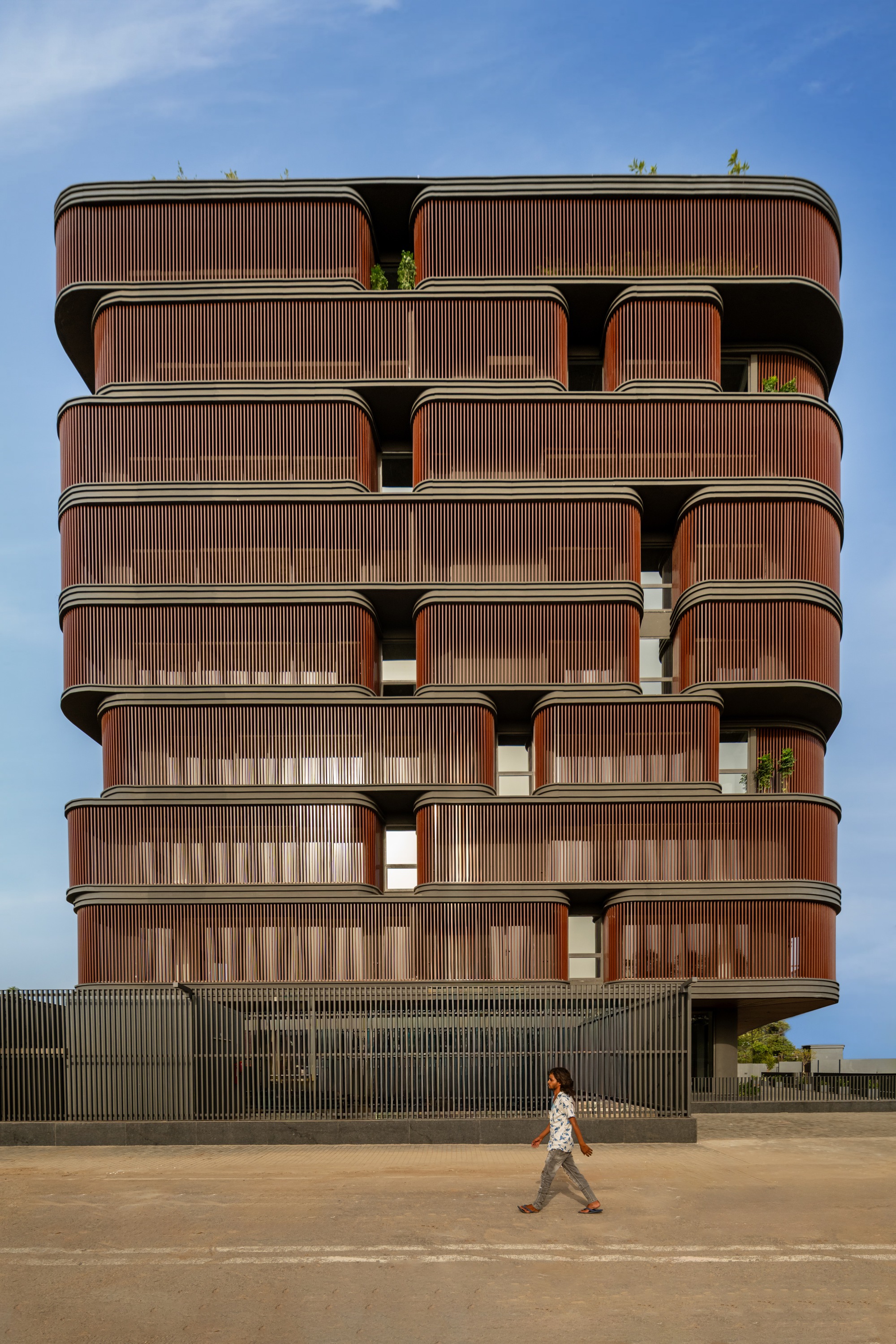 Stacked facade with vertical timber-slatted balconies and a person crossing the sandy forecourt below