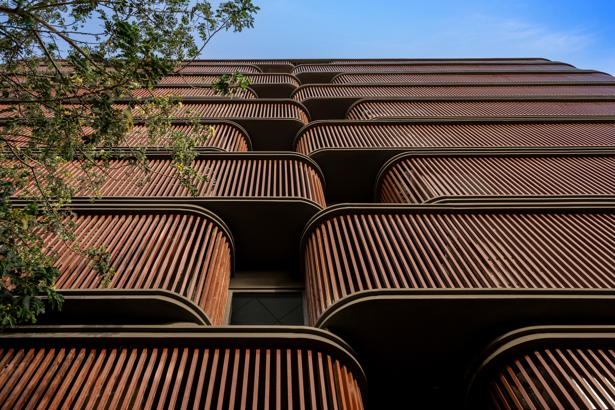 Facade with curved timber louvered balconies stacked vertically, framed by green foliage at left