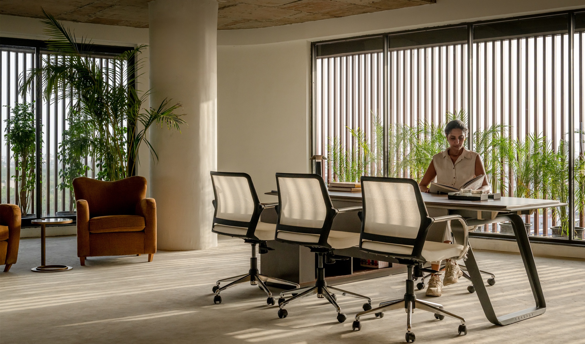 Interior workspace with exposed concrete ceiling, vertical louvered windows, potted palms and a person at a desk