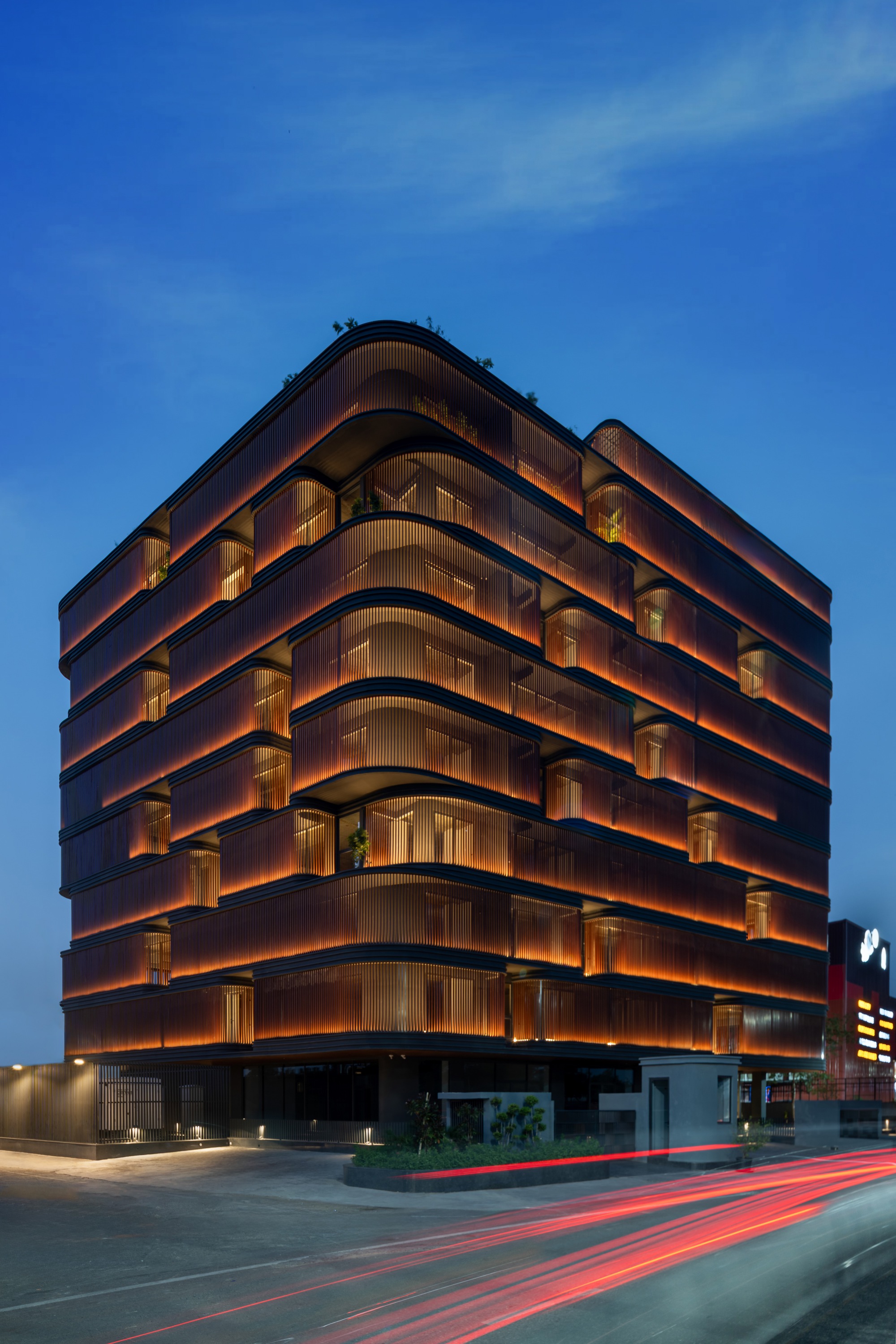Corner view of the illuminated facade with curved balconies and bronze louvers at dusk with traffic trails