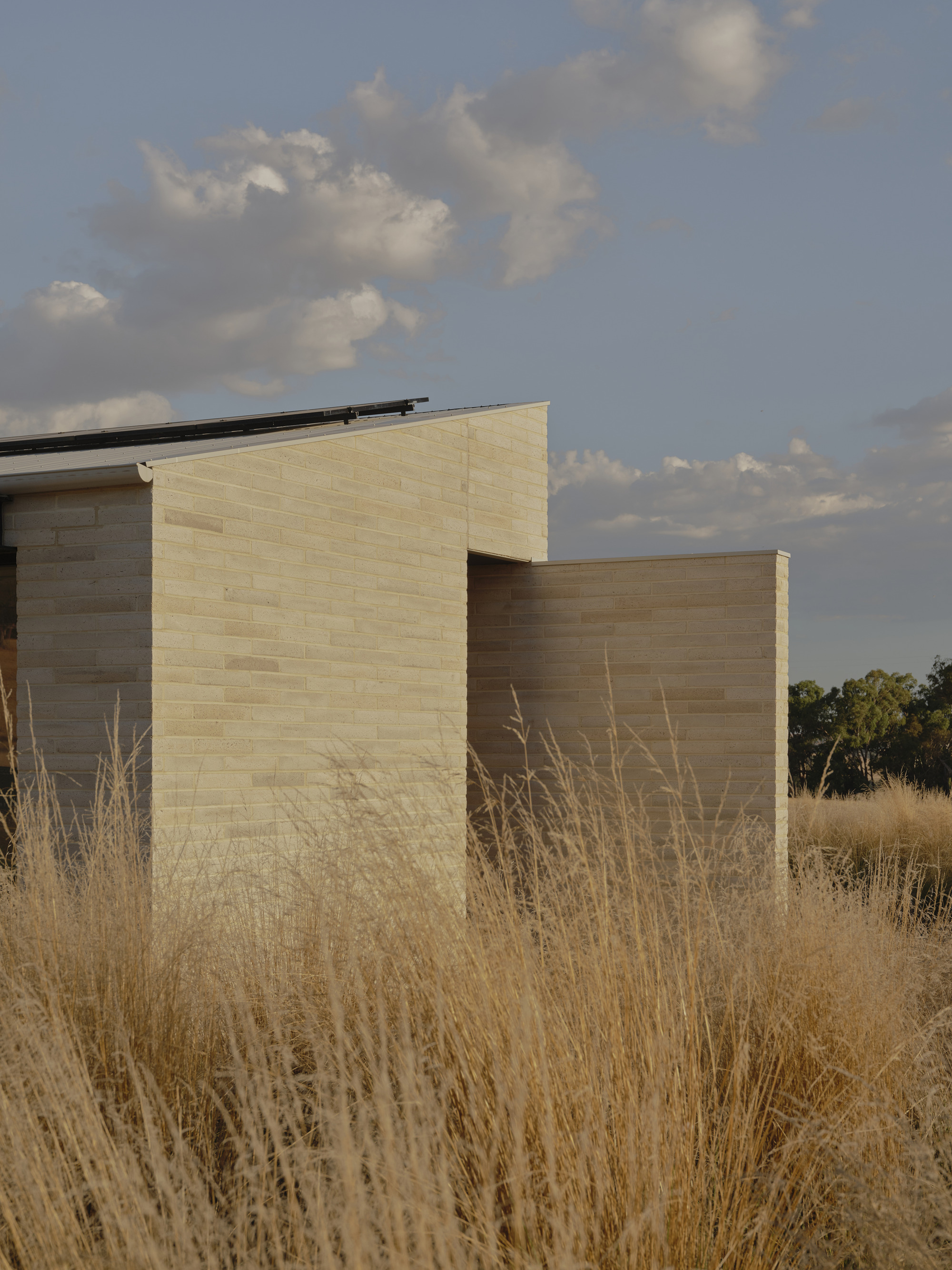 Two masonry volumes in contrasting tones emerging from dry grasses under a cloudy sky