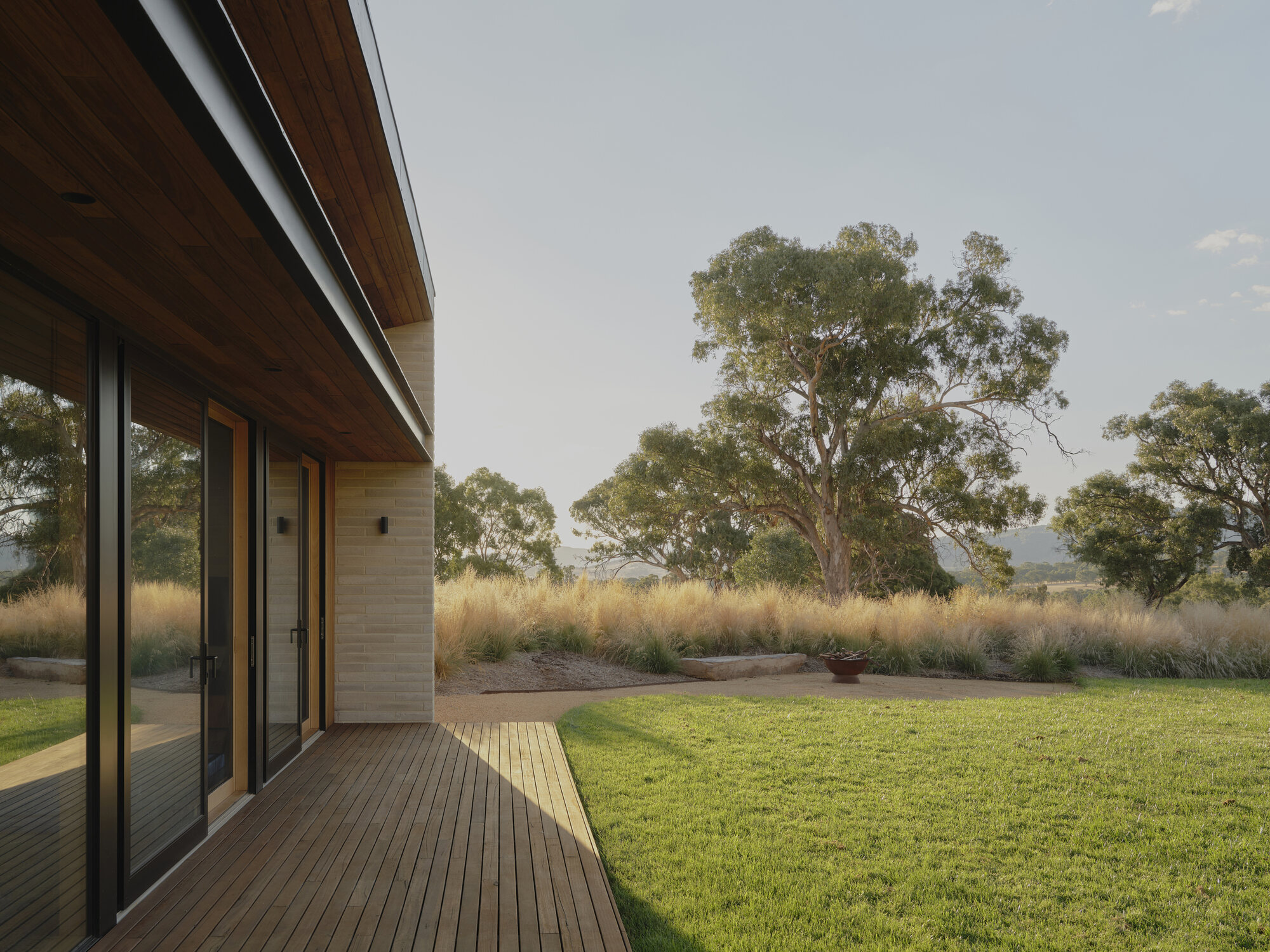 Timber deck extending from sliding glass doors toward a lawn and distant eucalyptus trees at dusk