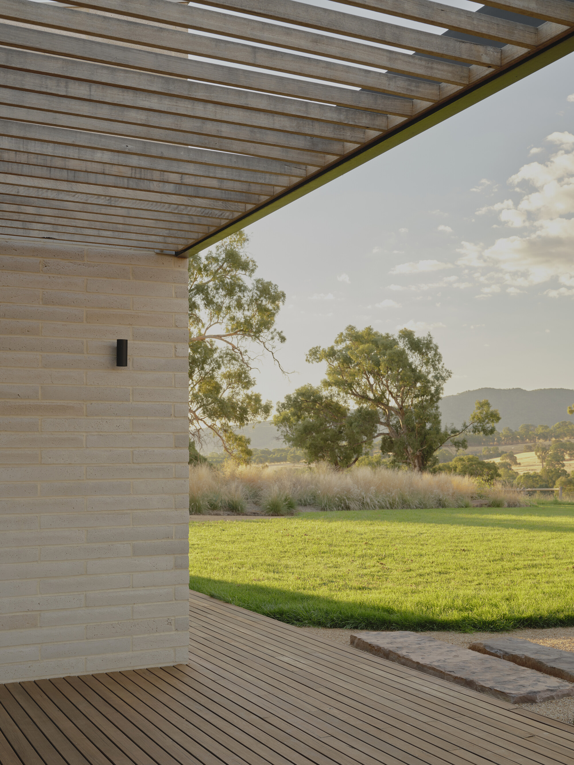 Corner of covered timber deck with slatted pergola overhead opening to lawn and distant trees