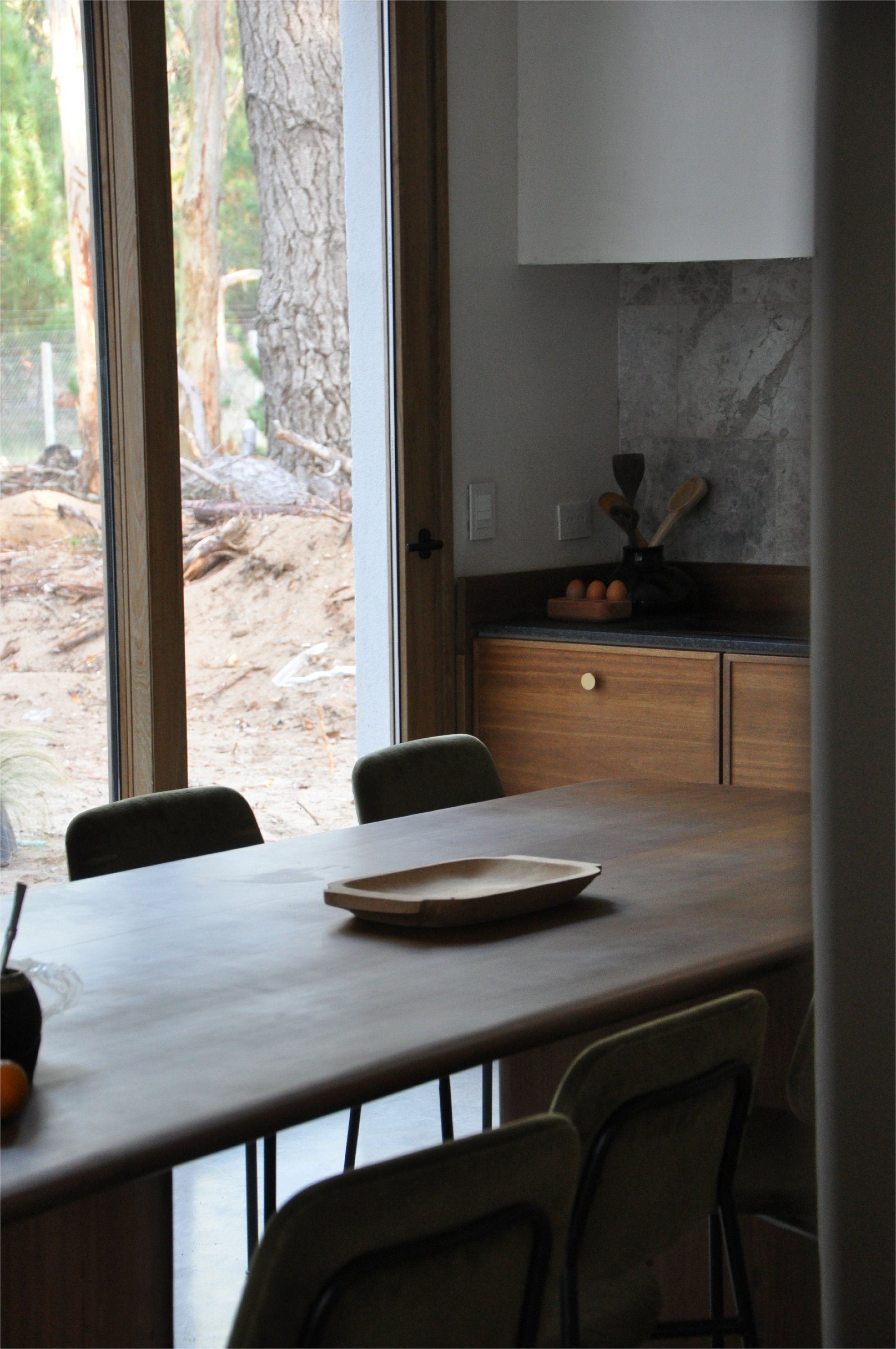 Dining table with timber kitchen cabinetry and grey stone backsplash beyond glazed doors