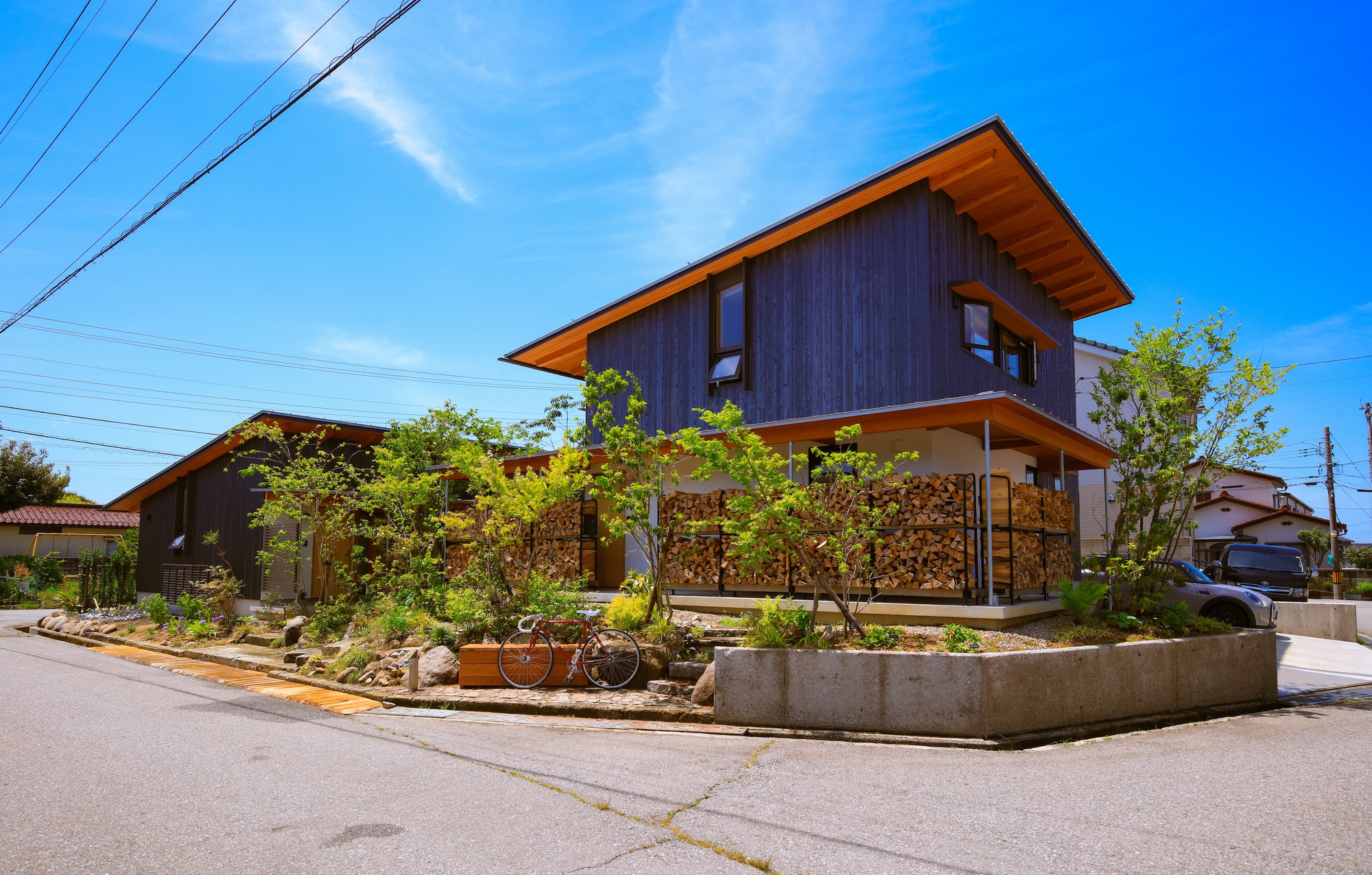 Street view of dark timber clad volumes with tilted roofs above a planted concrete curb
