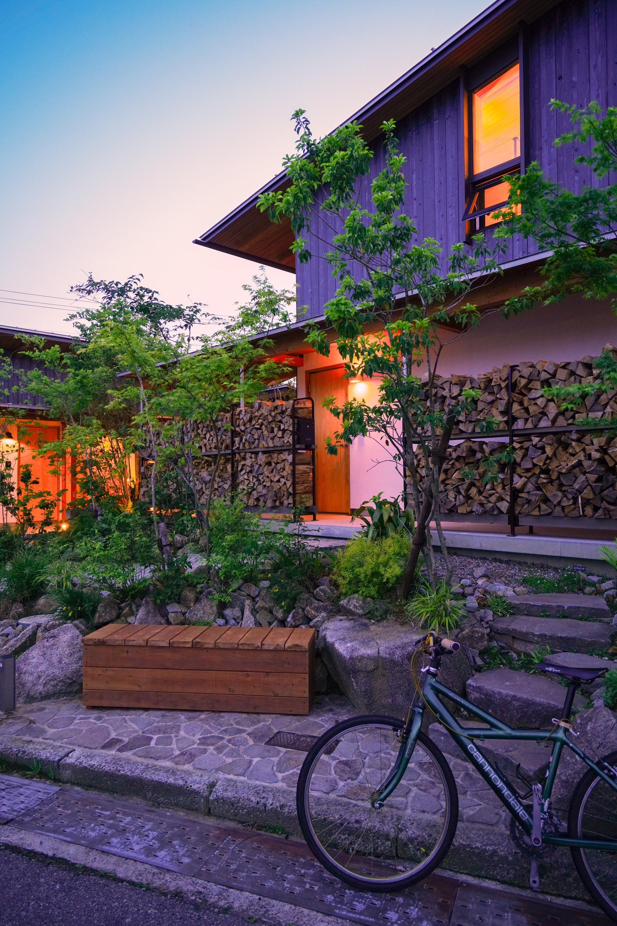 Front garden with planted beds, stacked firewood screen, and a bicycle at dusk
