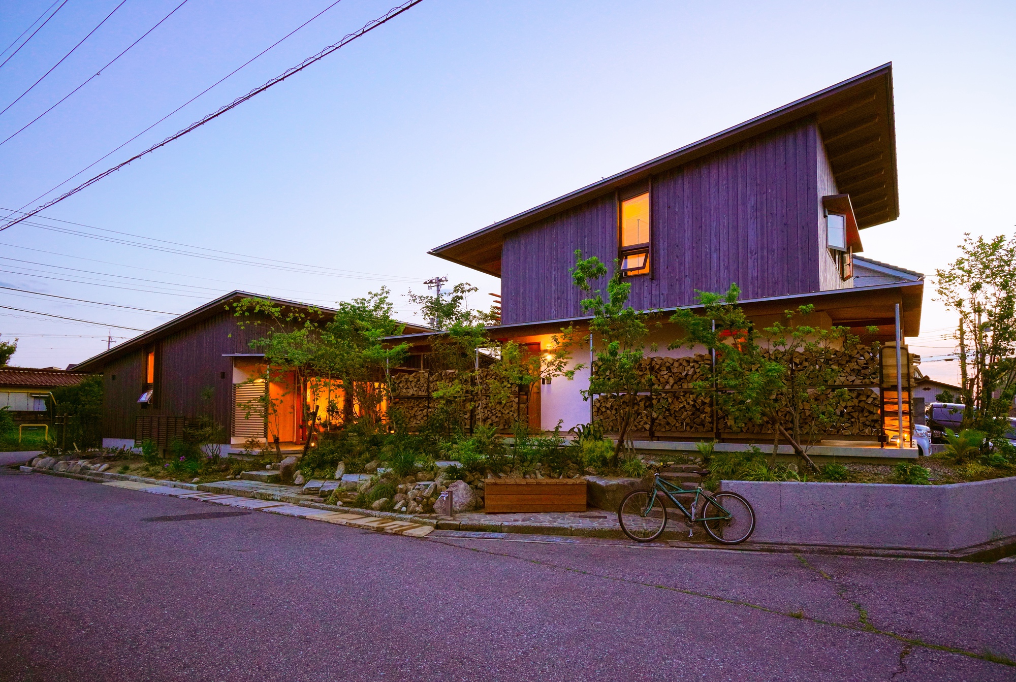 Street view of the vertical-clad volumes with planted garden beds and a bicycle at dusk