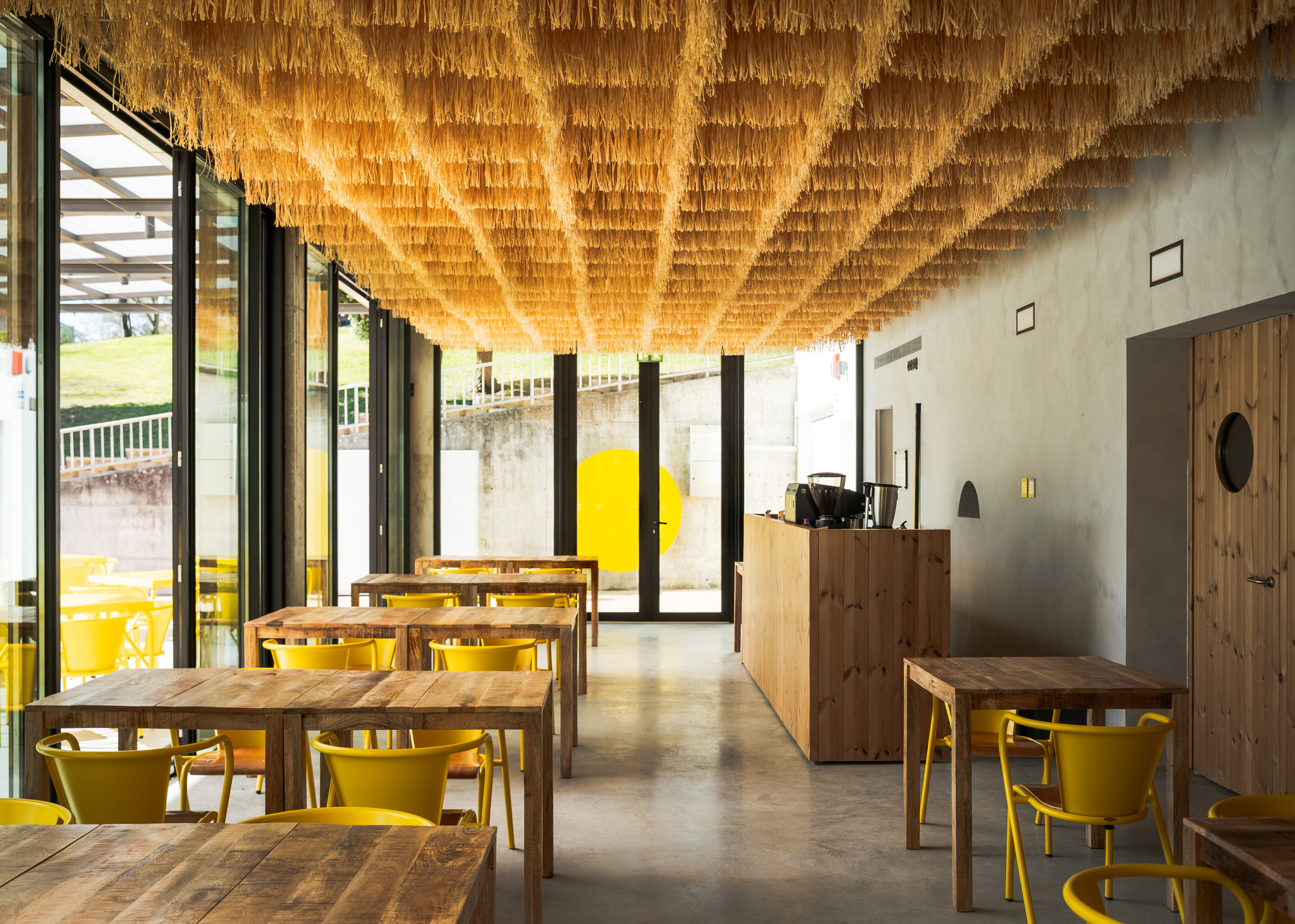 Dining room with layered raffia ceiling installation and yellow chairs beside floor-to-ceiling glazing