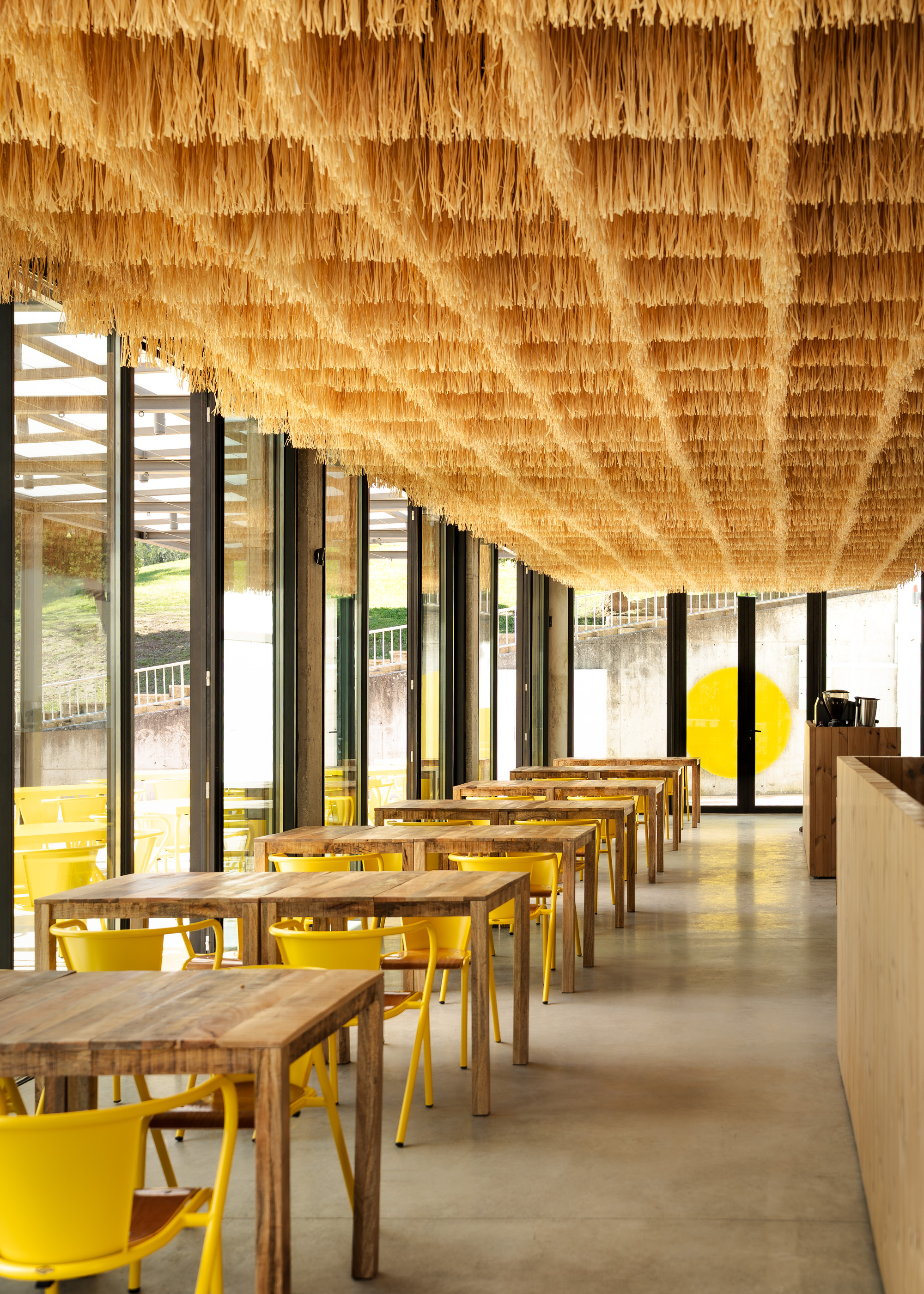 Dining space with layered palm frond ceiling installation above timber tables and yellow chairs