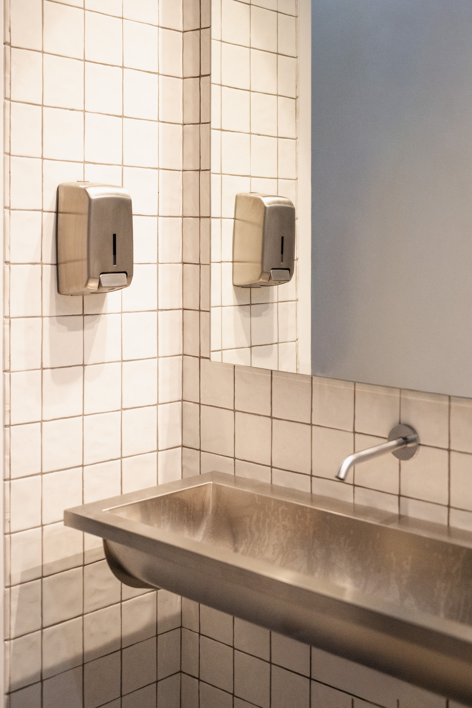 Corner washroom detail with white square tile and wall-mounted soap dispensers above a limestone trough sink