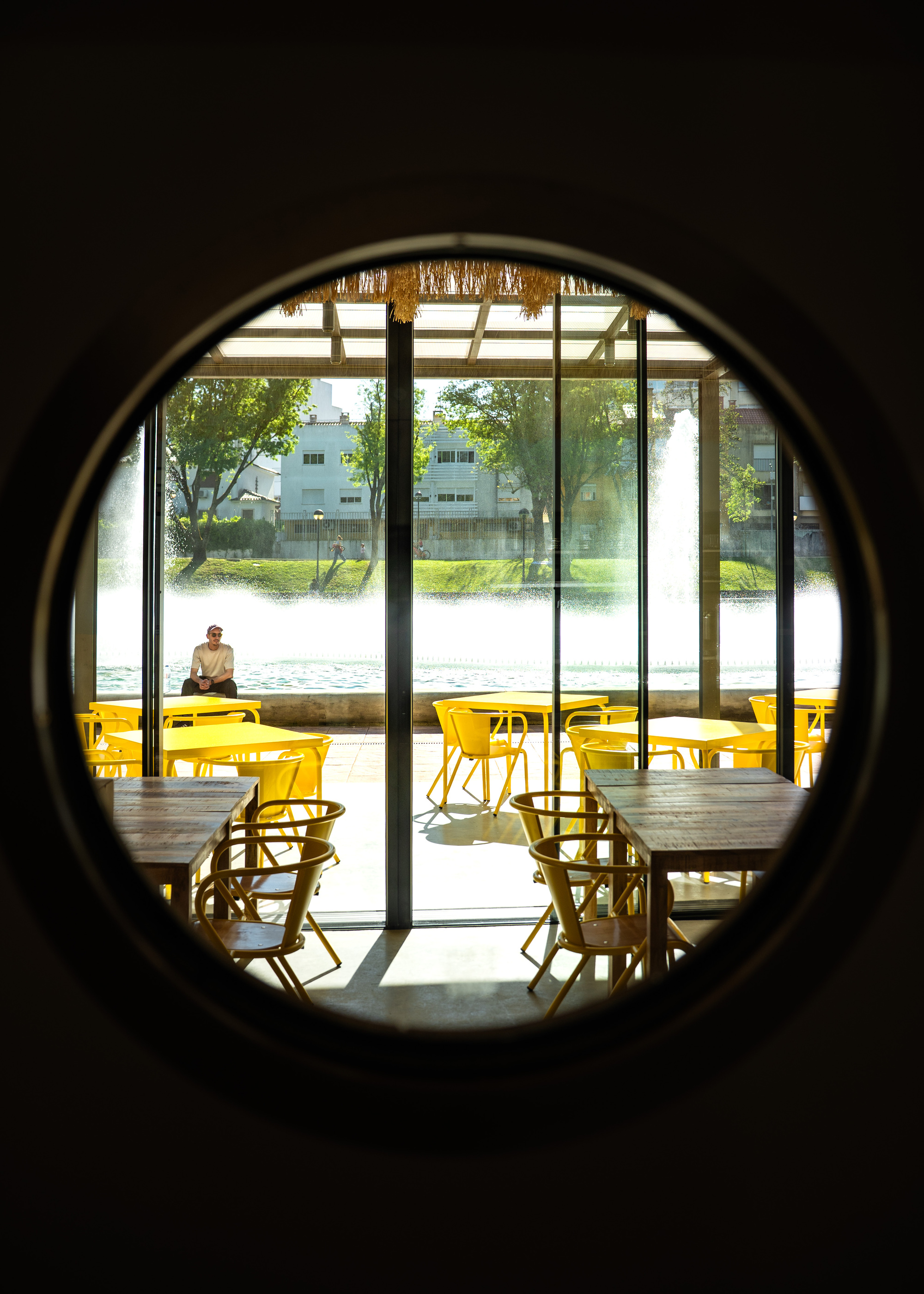 View through circular portal opening to dining area with yellow seating and glazed facade beyond