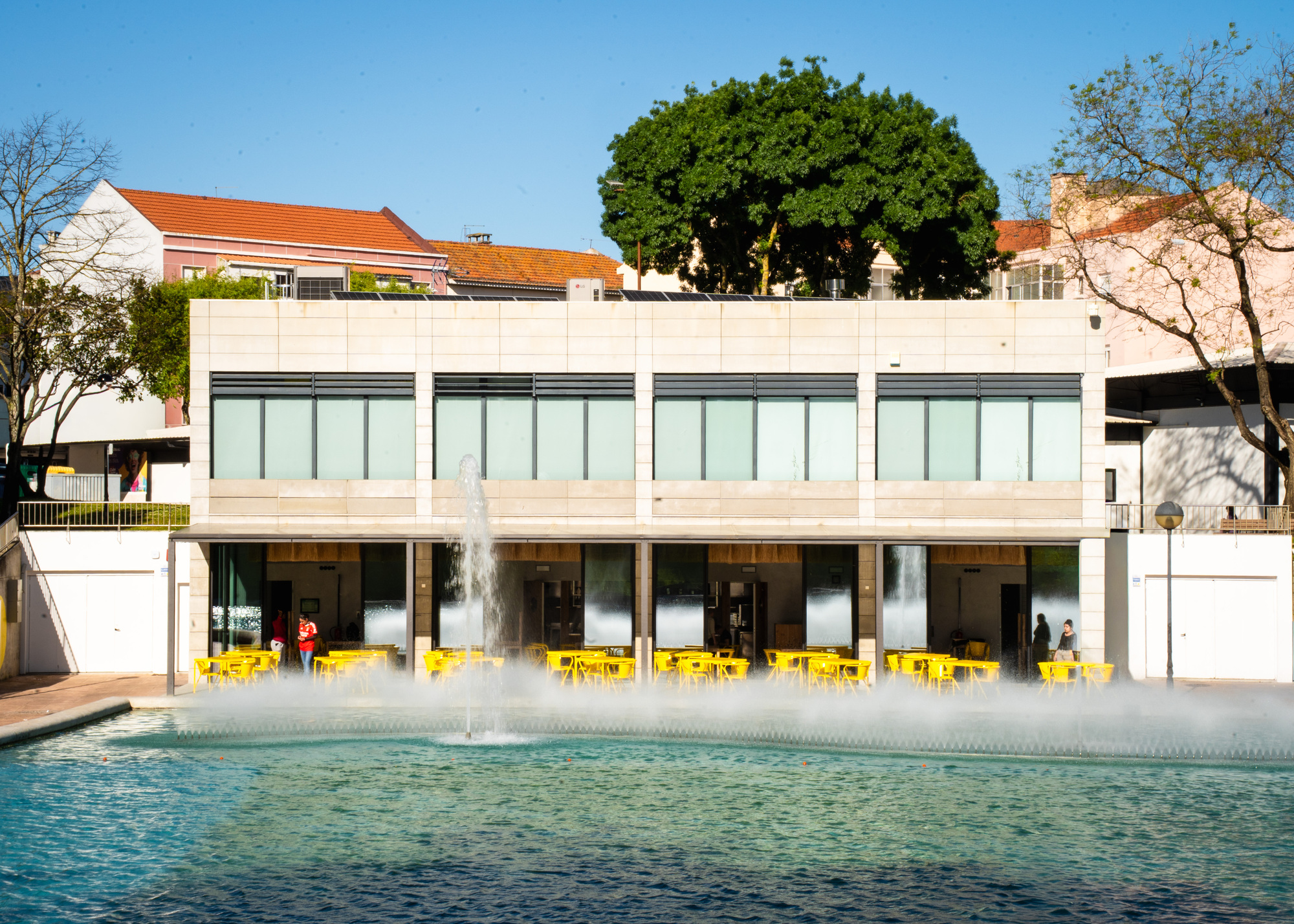 Two-story facade with upper glass curtain wall and lower colonnade opening onto pool with fountain jets