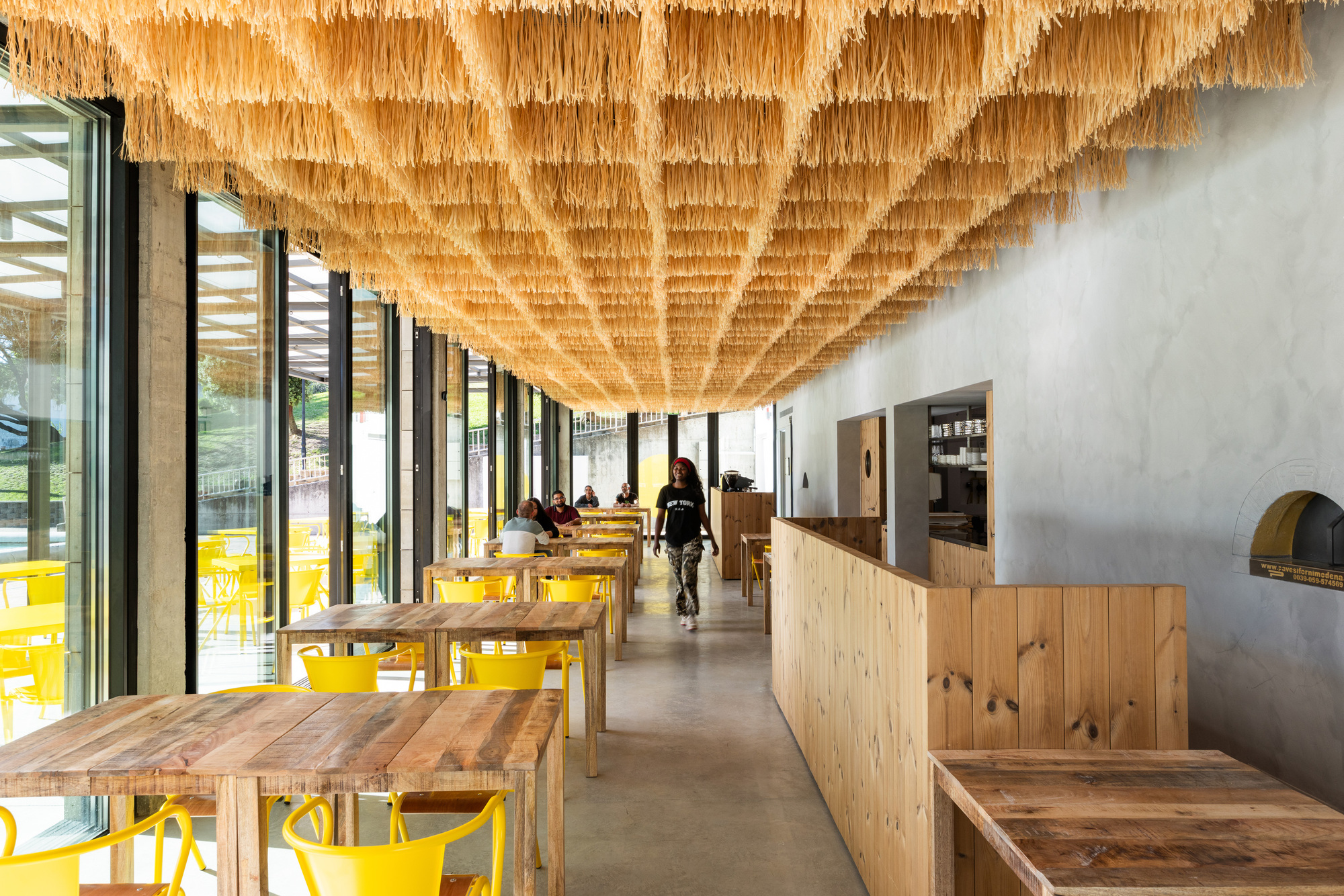 Dining hall with timber tables, yellow chairs, and hanging woven ceiling installation over a polished concrete floor