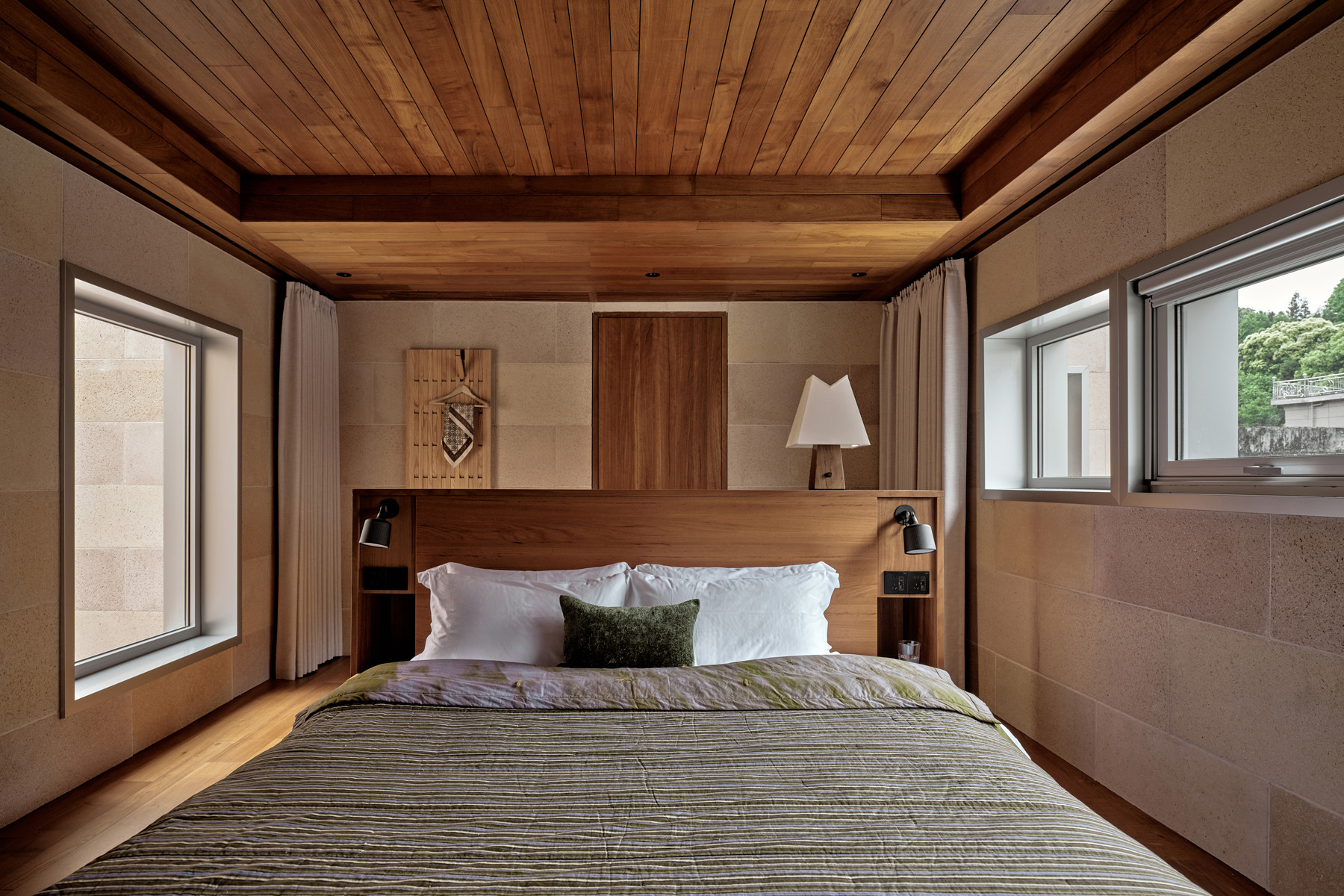Bedroom with timber headboard and ceiling between sandstone walls with windows on each side