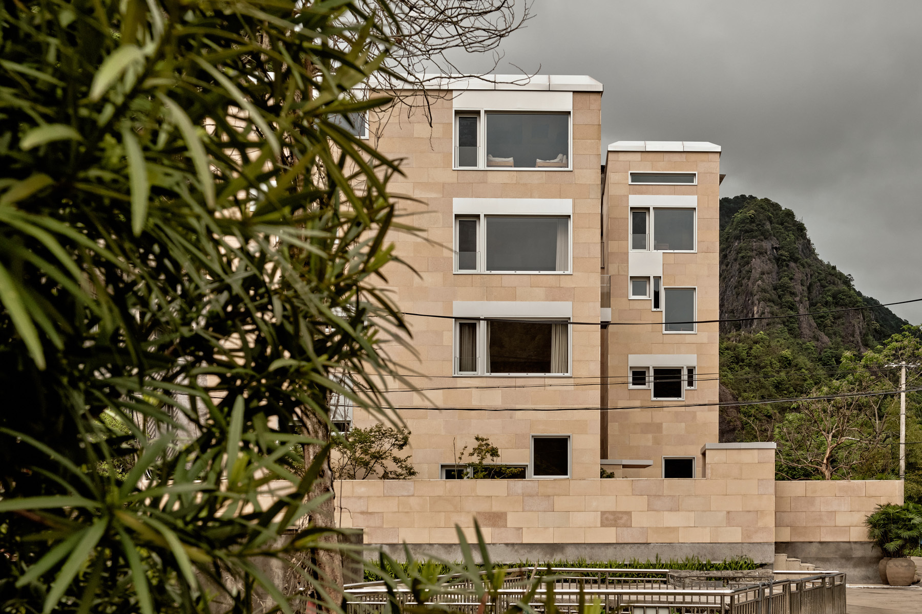 Facade of stacked block volumes with punched windows framed by foreground foliage and a volcanic peak behind
