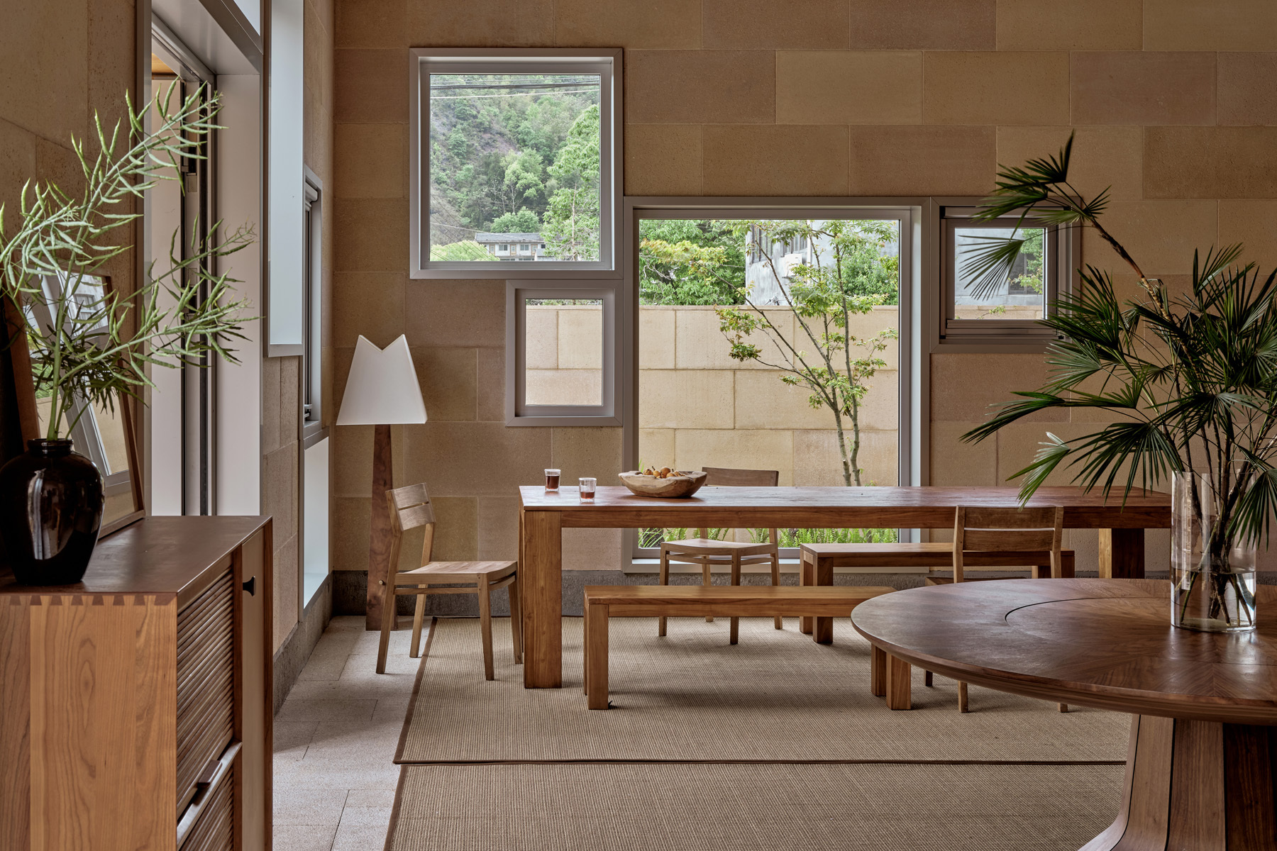 Dining area with timber table and benches beneath scattered square windows overlooking greenery and neighboring walls