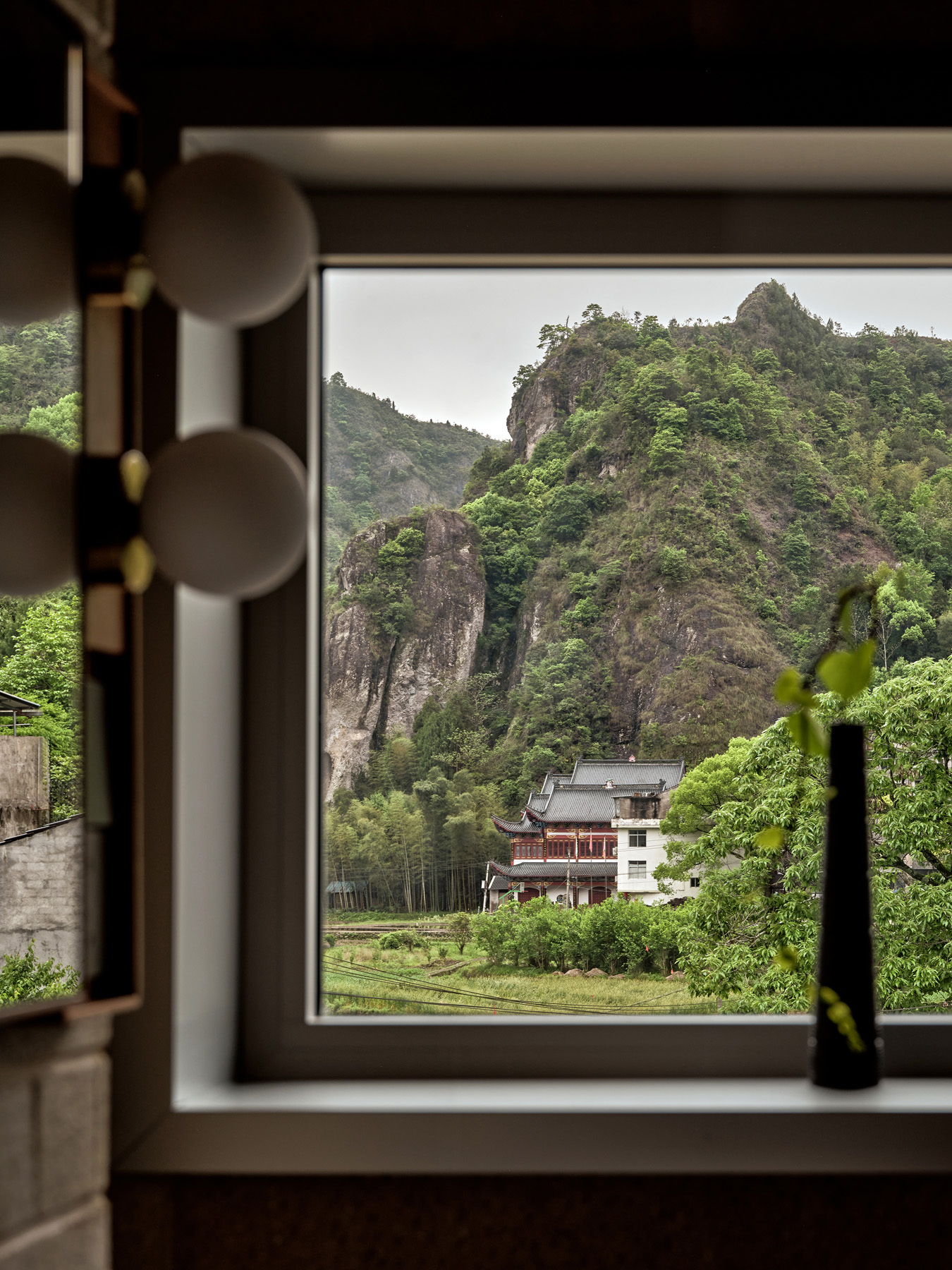 Window framing a view of forested karst mountains and a neighboring building in the valley