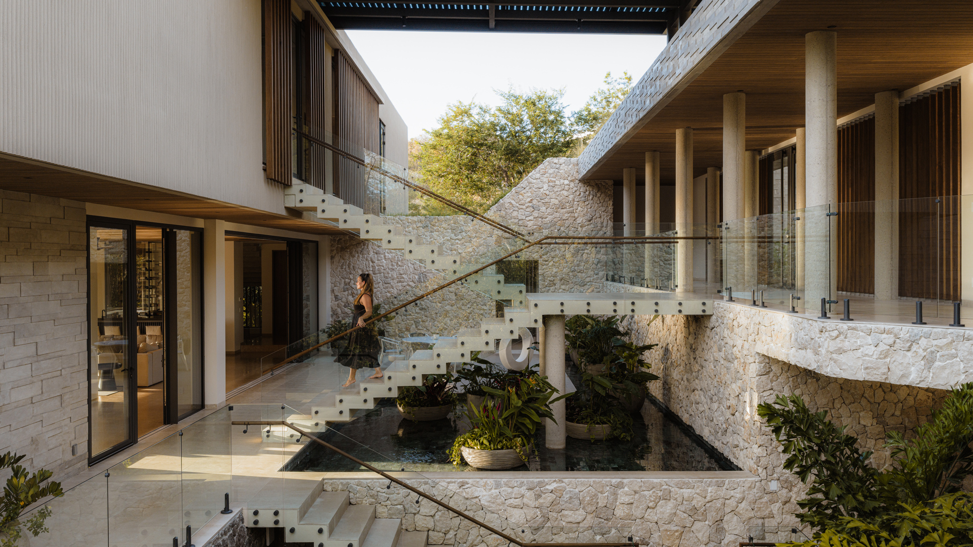 Glass staircase spanning courtyard pond between stone-clad volumes with timber ceiling overhead