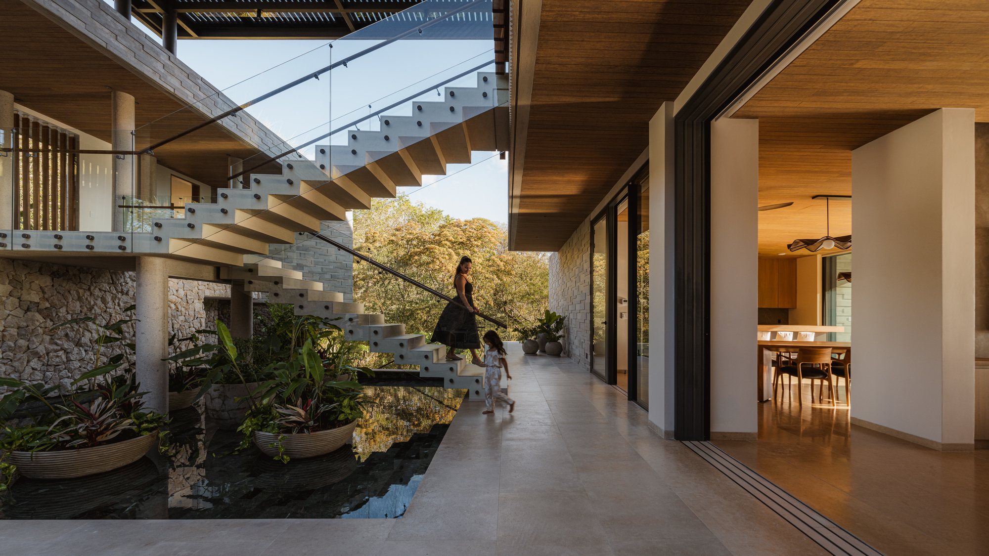 Open-air corridor with floating staircase, water feature, and a woman with child in evening light