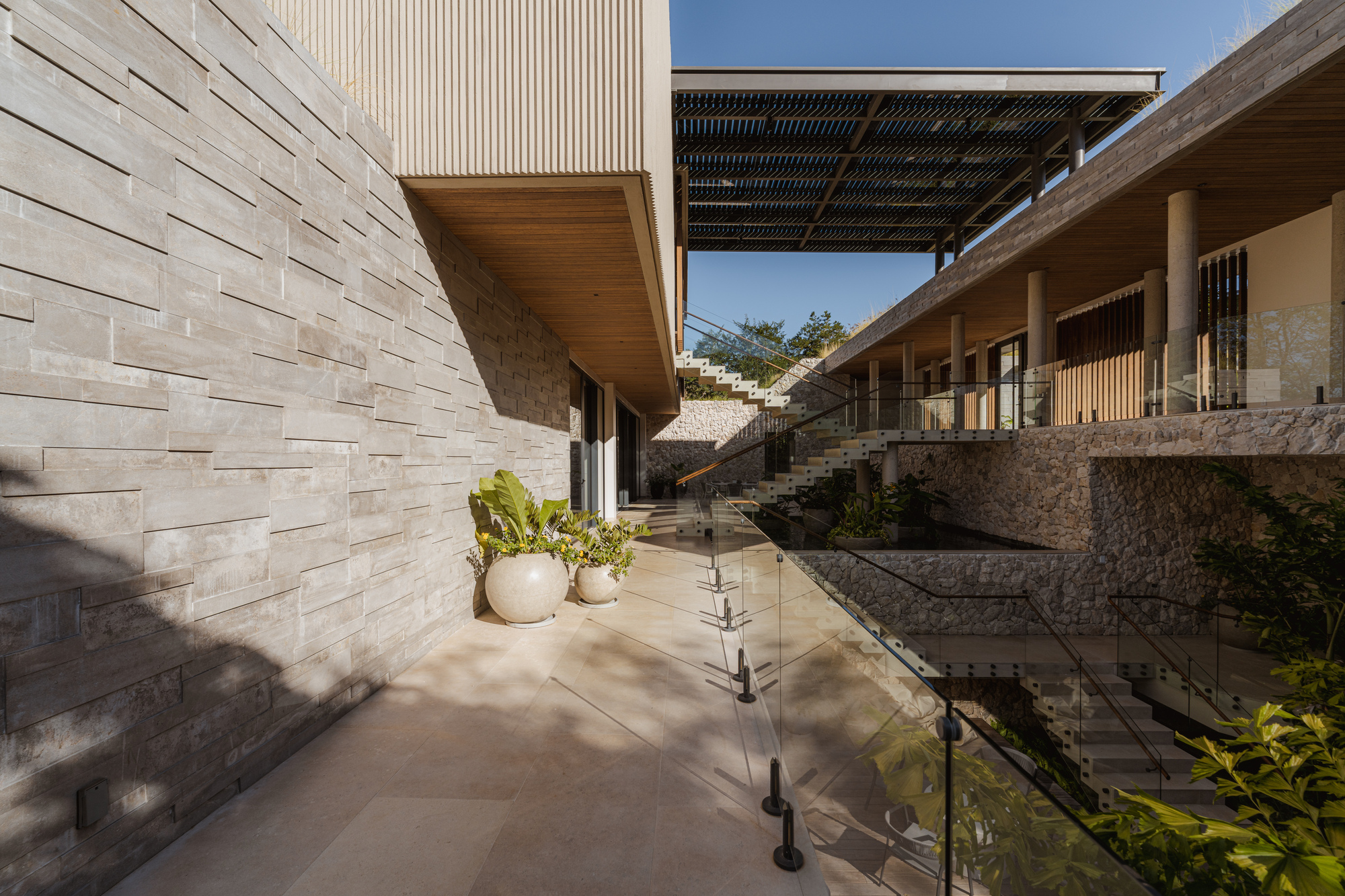 Outdoor terrace with textured stone wall and glass balustrade overlooking planted steps and perforated canopy