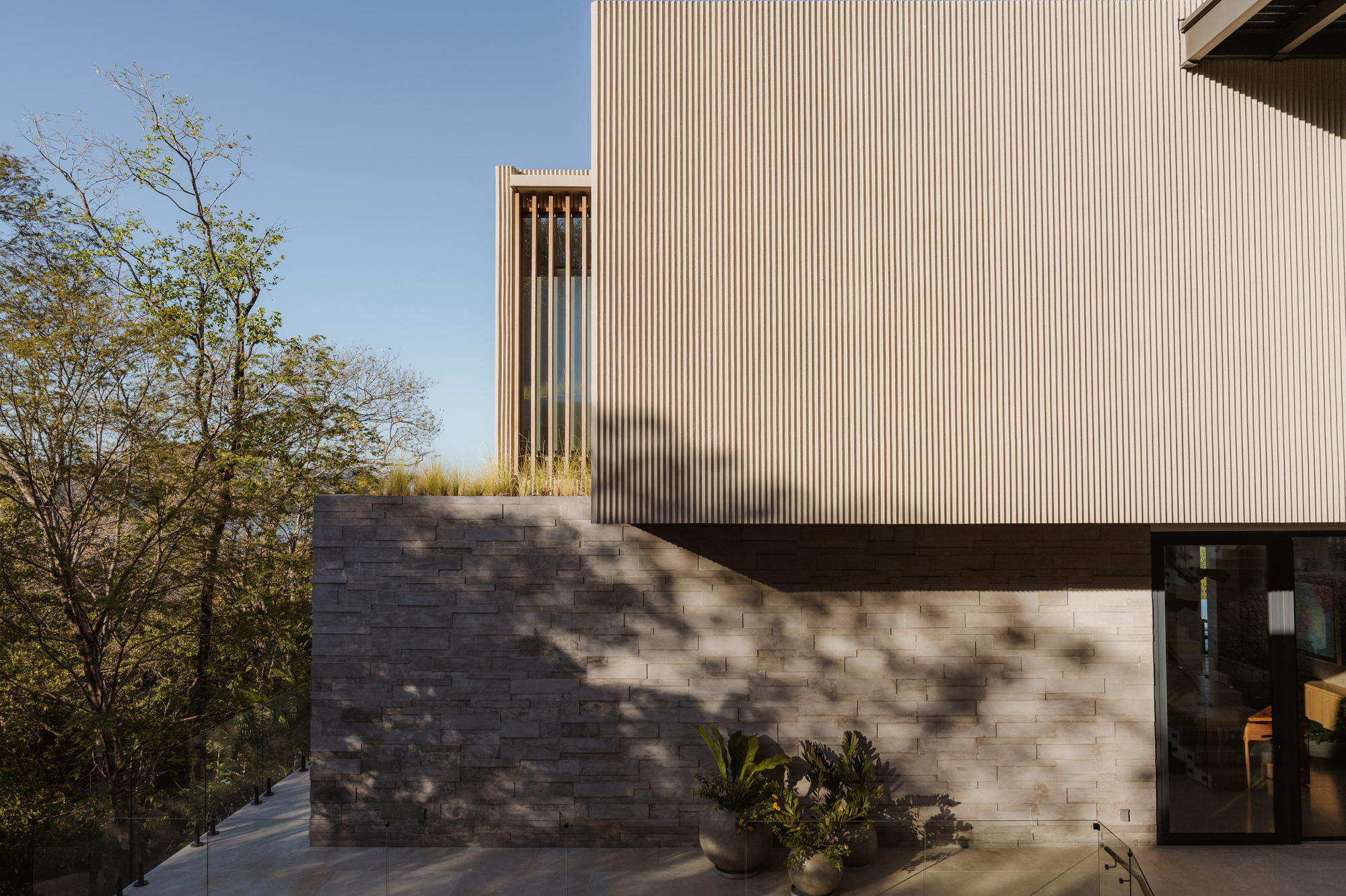 Exterior corner showing stacked stone base supporting vertical timber slat cladding with planted foreground