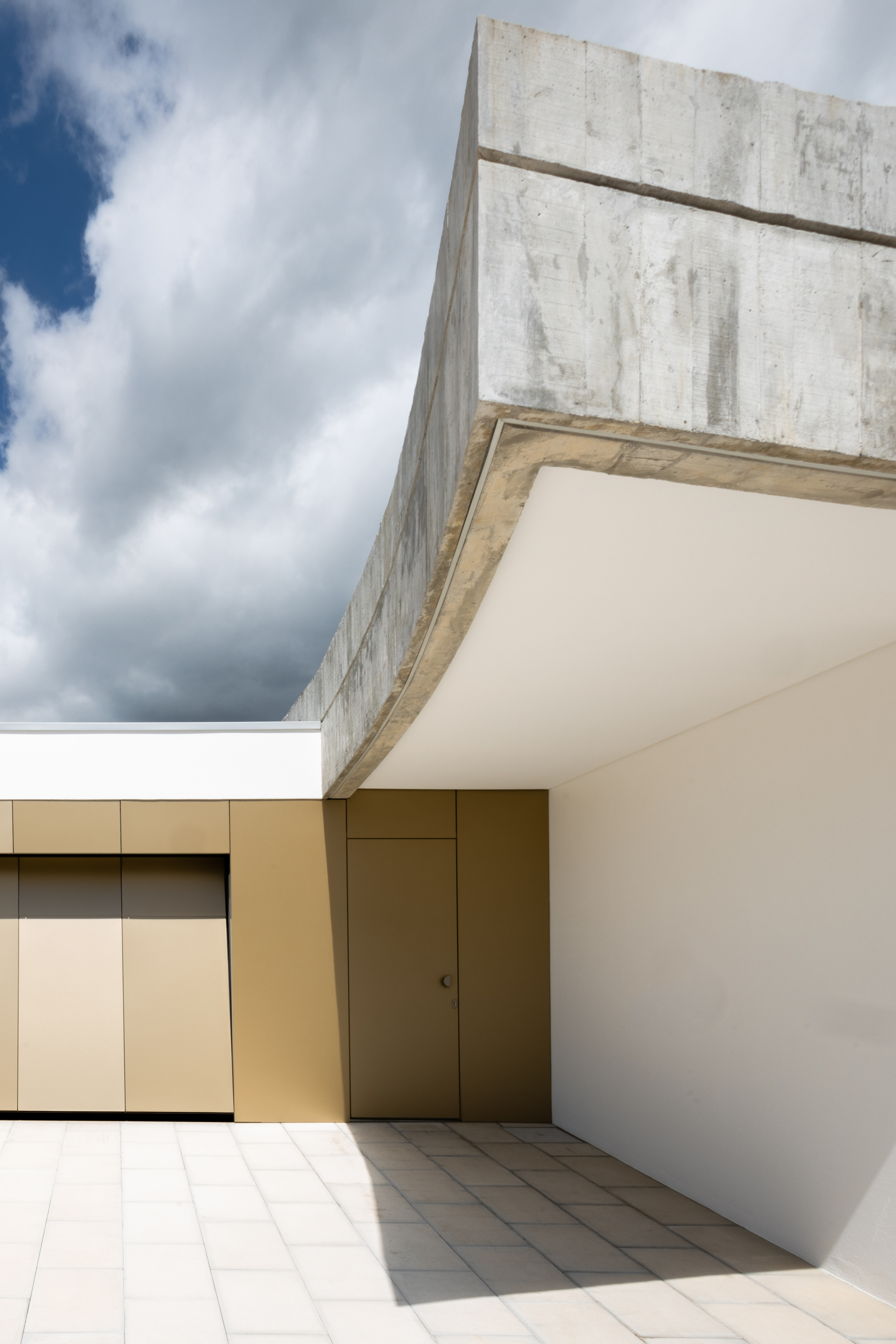 Courtyard corner with board-formed concrete parapet and cream-rendered walls under a cloudy sky