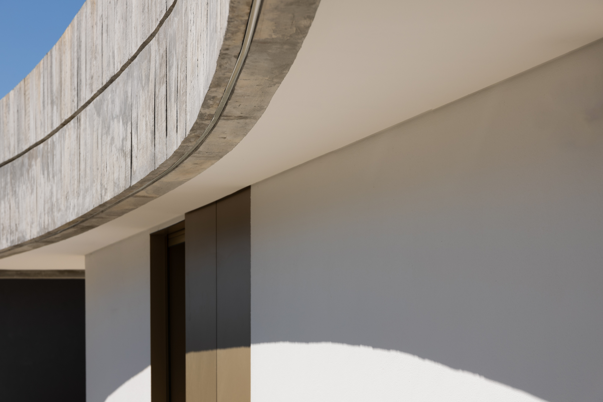 Detail of curved board-formed concrete parapet edge casting shadows on cream rendered wall