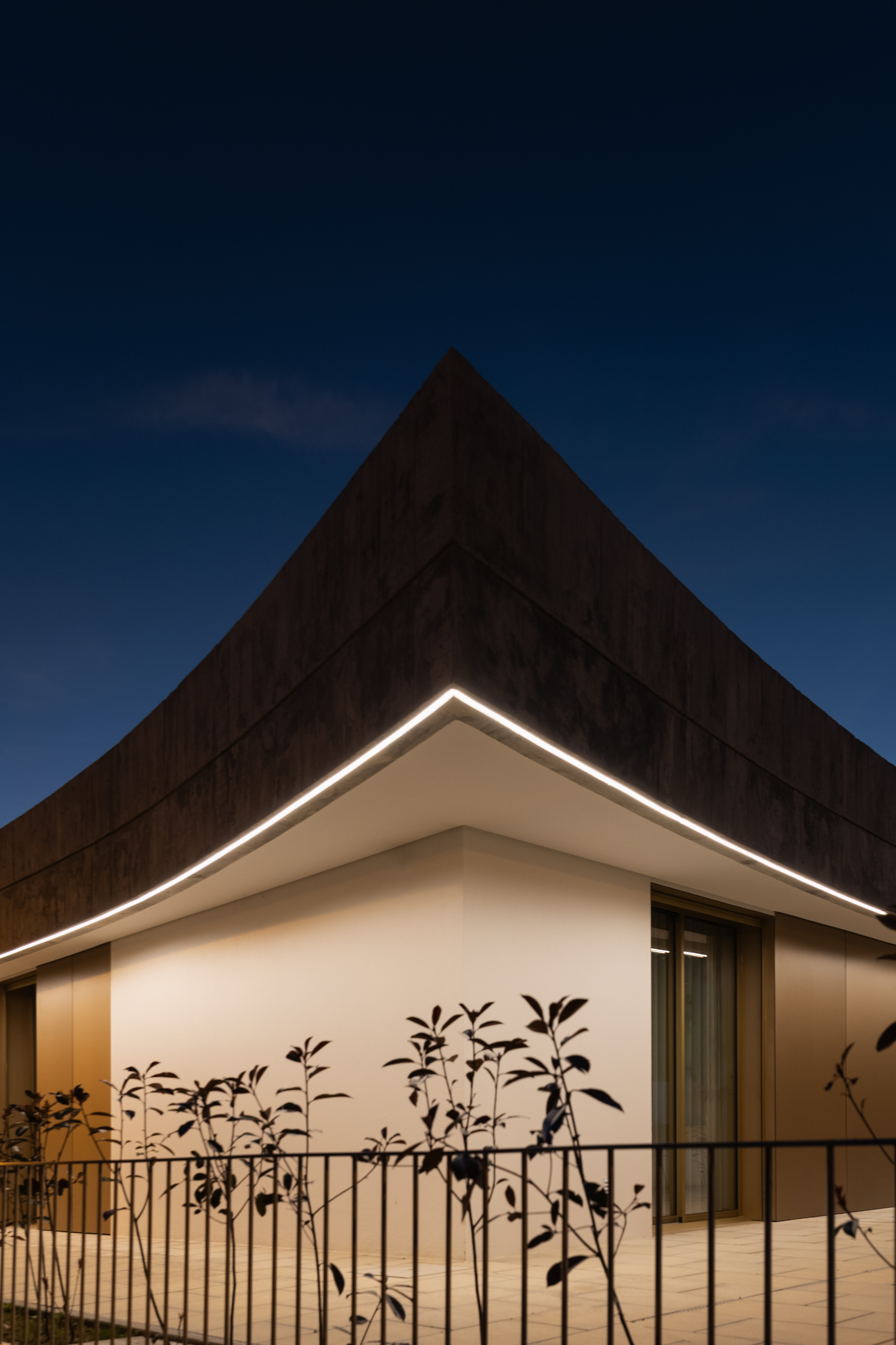 Illuminated roof edge above the white facade corner with bamboo plants behind a metal railing