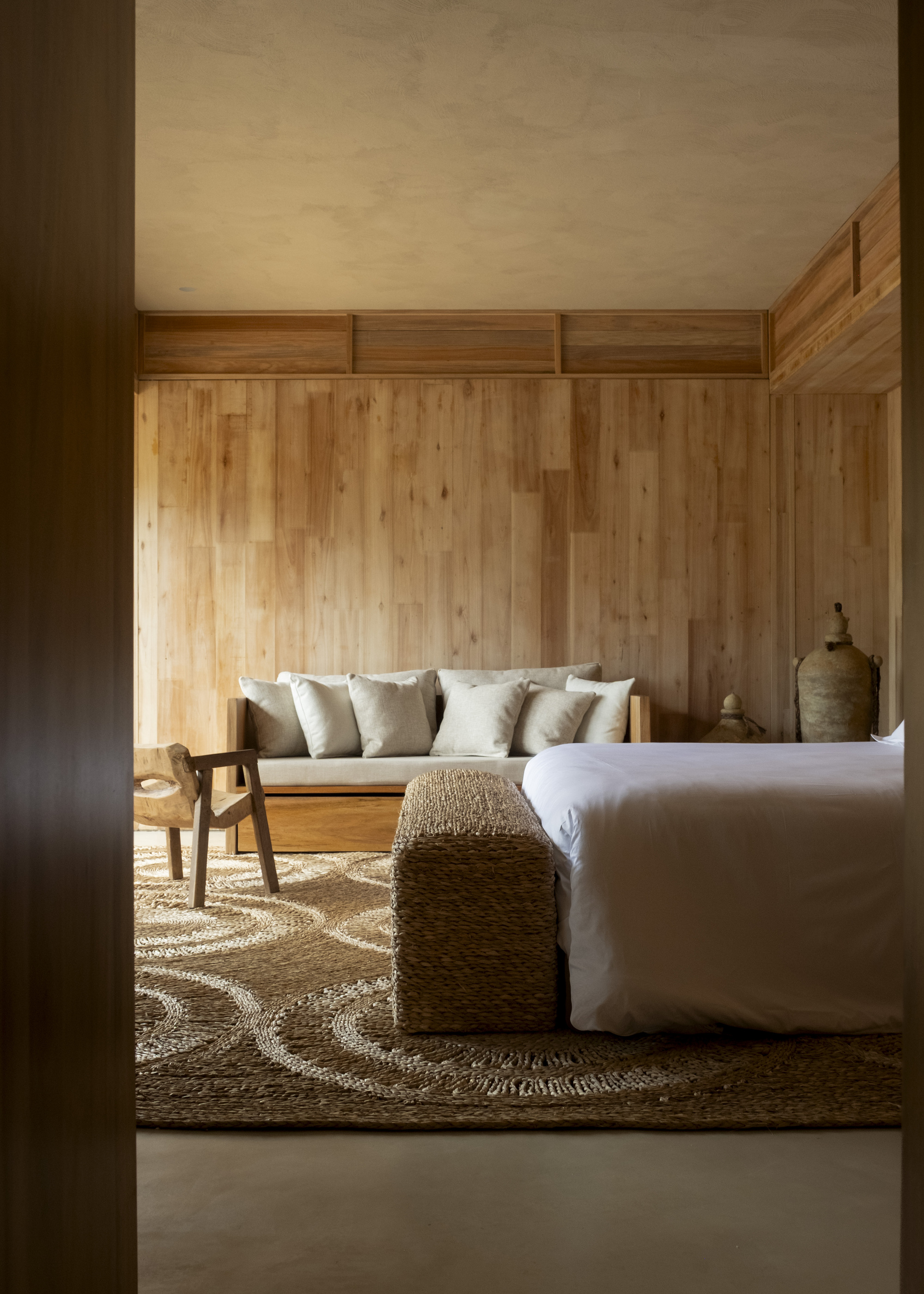 Bedroom interior with vertical timber plank walls and transom windows filtering afternoon light across a jute rug