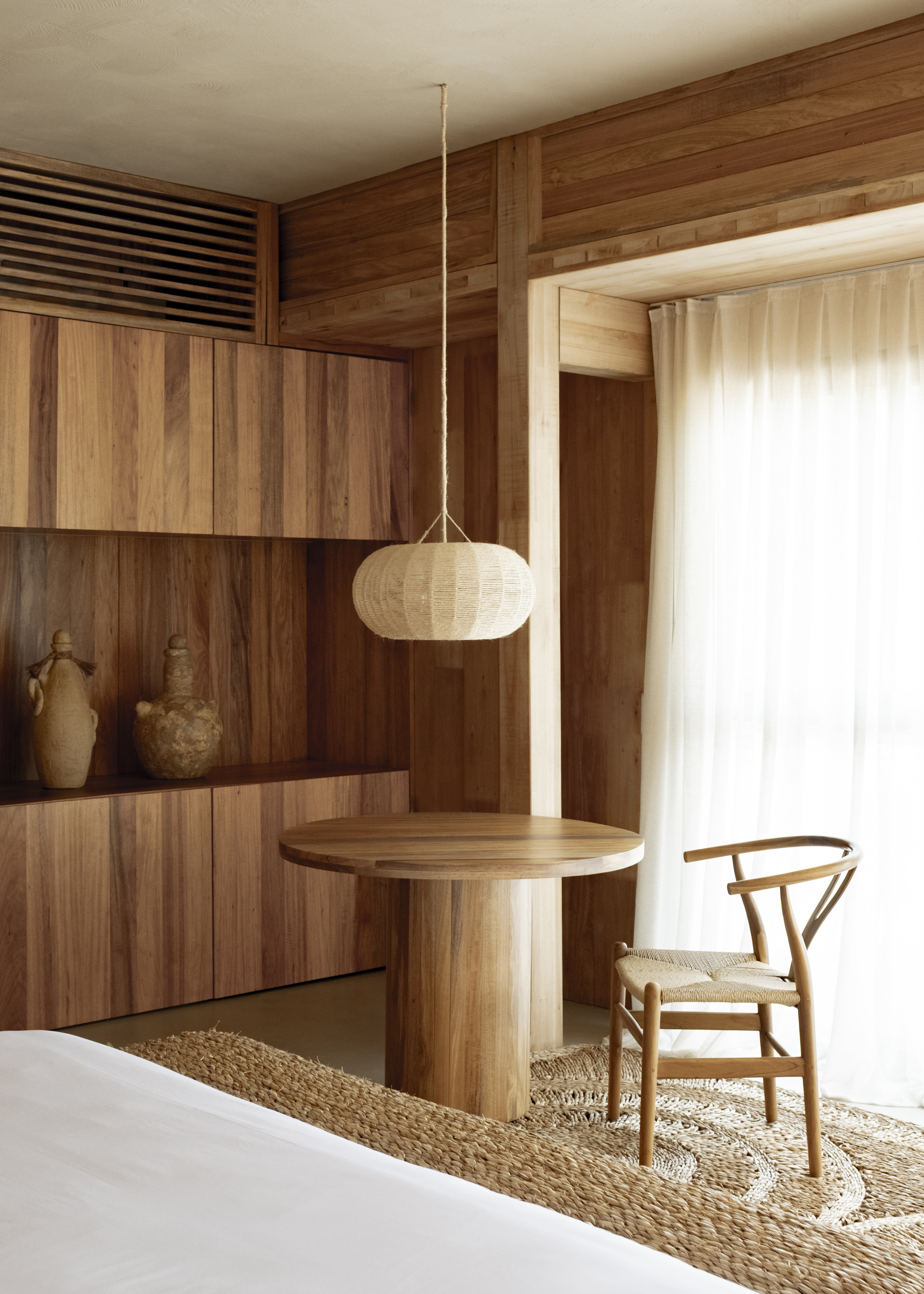 Interior corner with timber-paneled walls, built-in shelving, pendant lamp, and woven rug below a white curtain