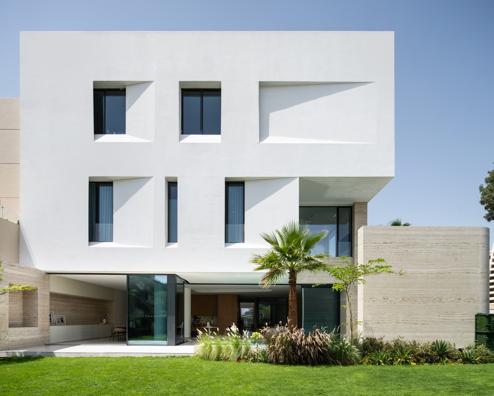 Garden facade showing cantilevered white volumes above travertine base and planted lawn with palm