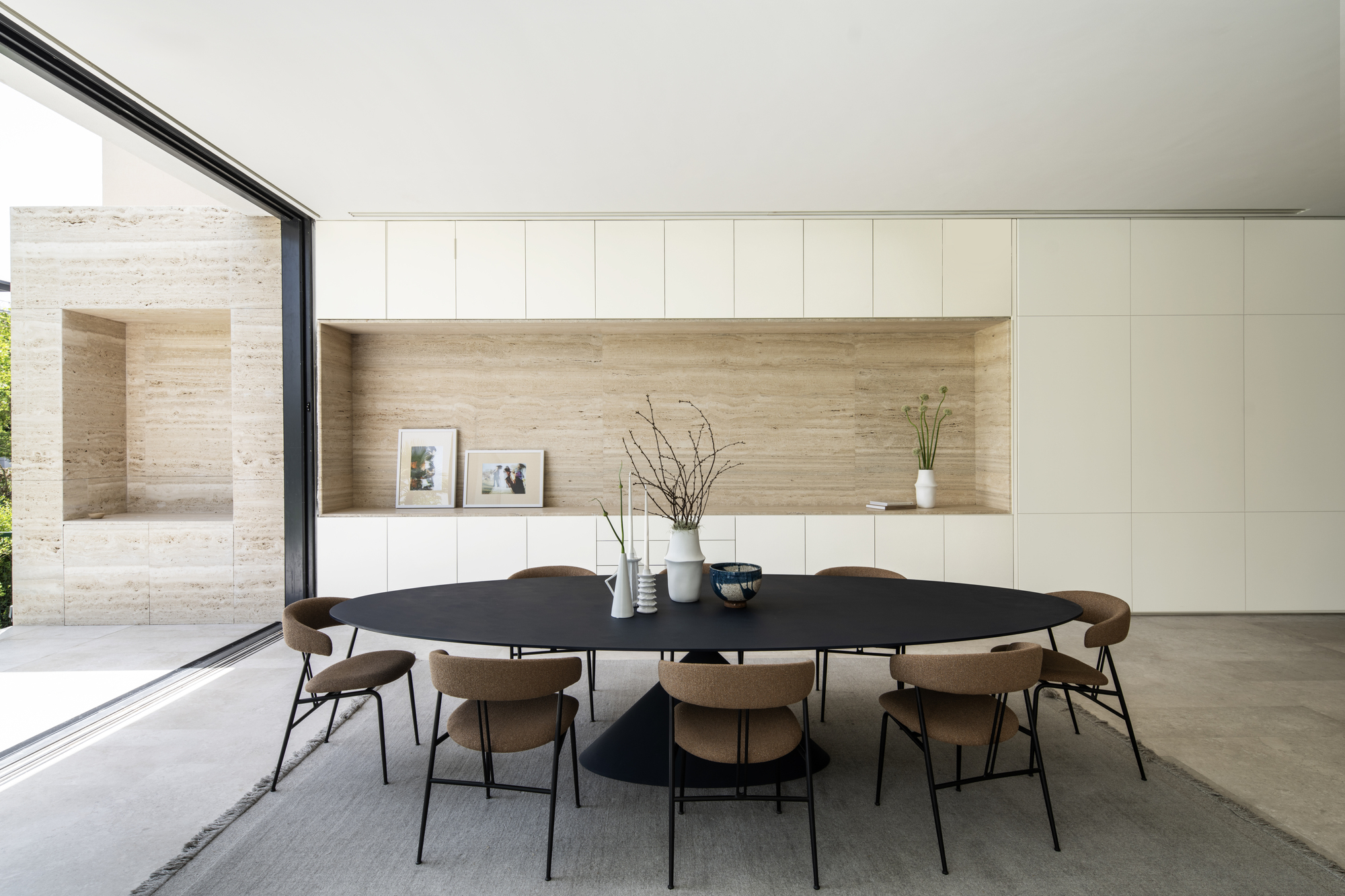 Dining area with dark oval table beneath white ceiling and horizontal travertine niche in pale cabinetry