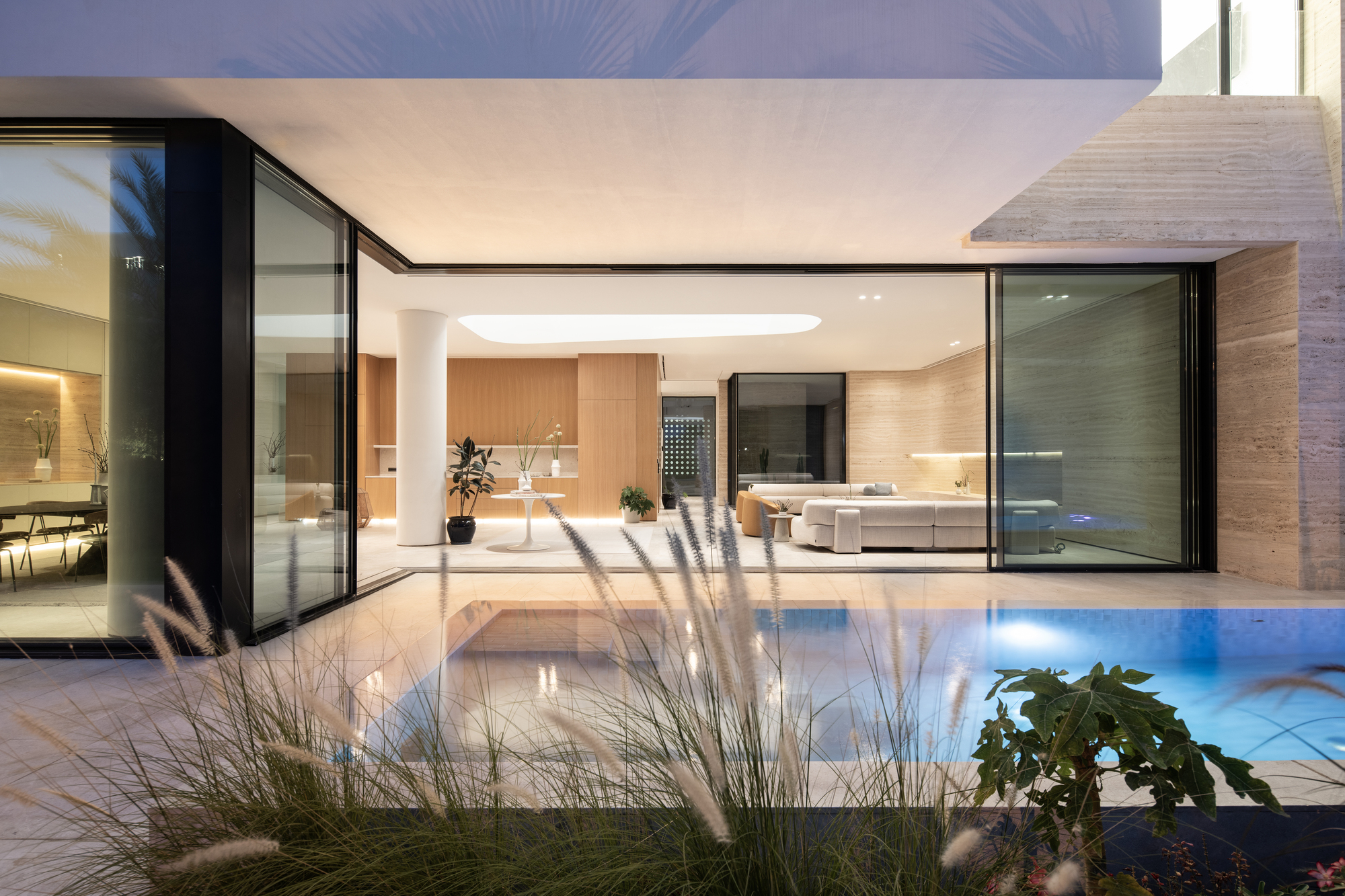 Glazed living pavilion opening to illuminated pool with ornamental grasses in foreground at dusk