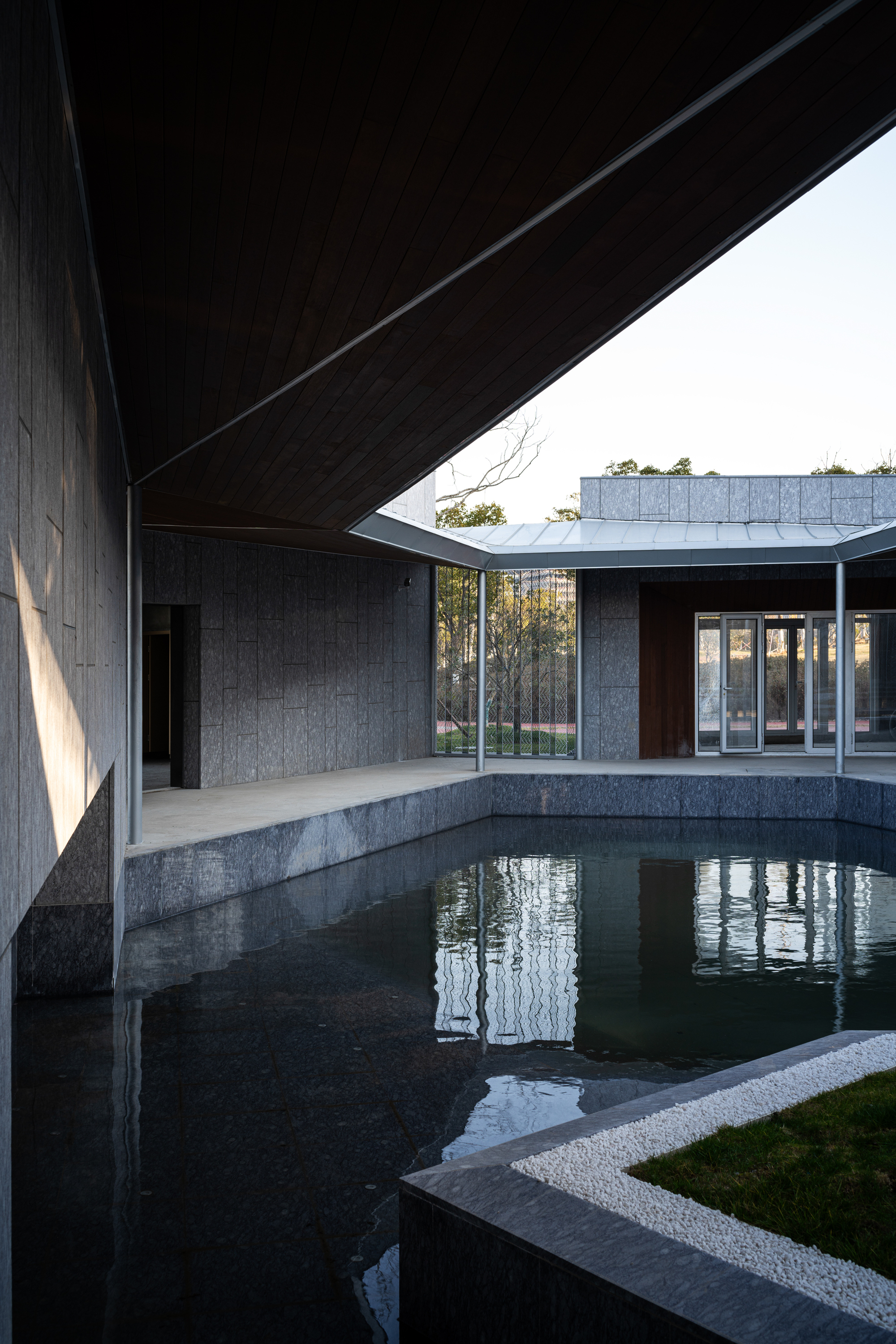 Covered walkway beside concrete-edged reflecting pool with charred timber cladding in afternoon shadow