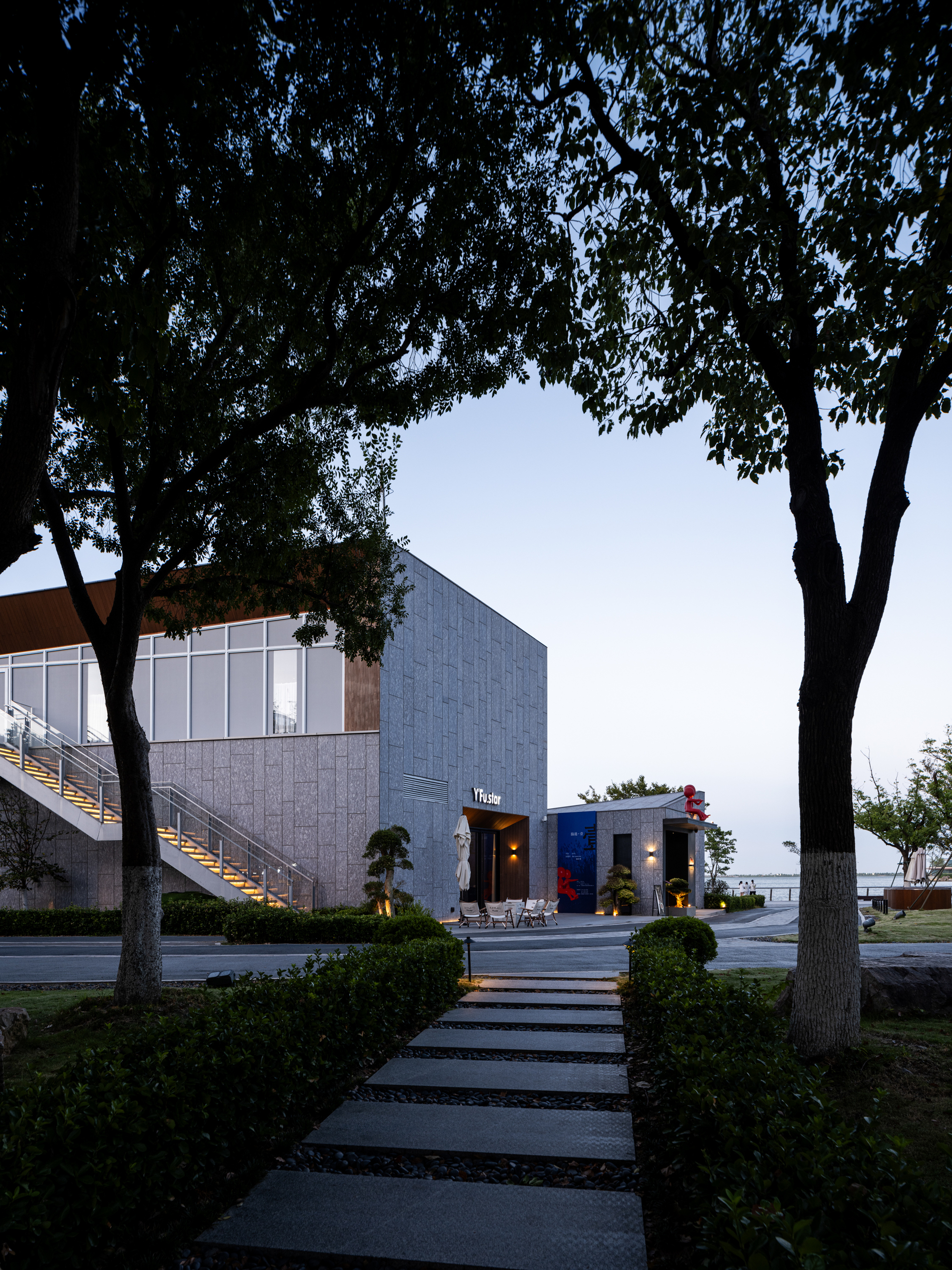Concrete paver pathway leading through mature trees toward cubic volumes at twilight