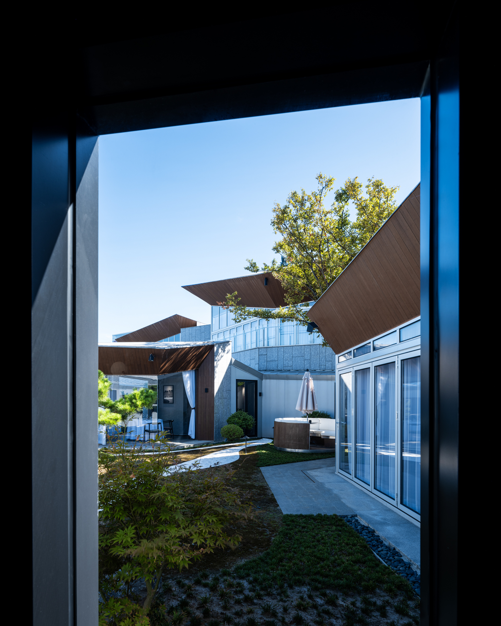 View through a doorway into a courtyard with corrugated metal volumes and angled timber canopies under clear sky