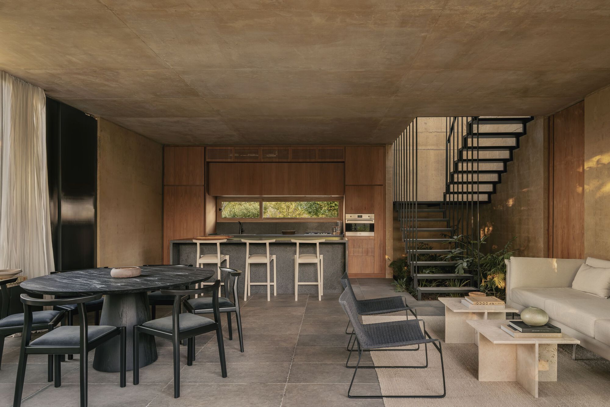 Open-plan living space with exposed concrete ceiling, timber kitchen and metal staircase beside dining area